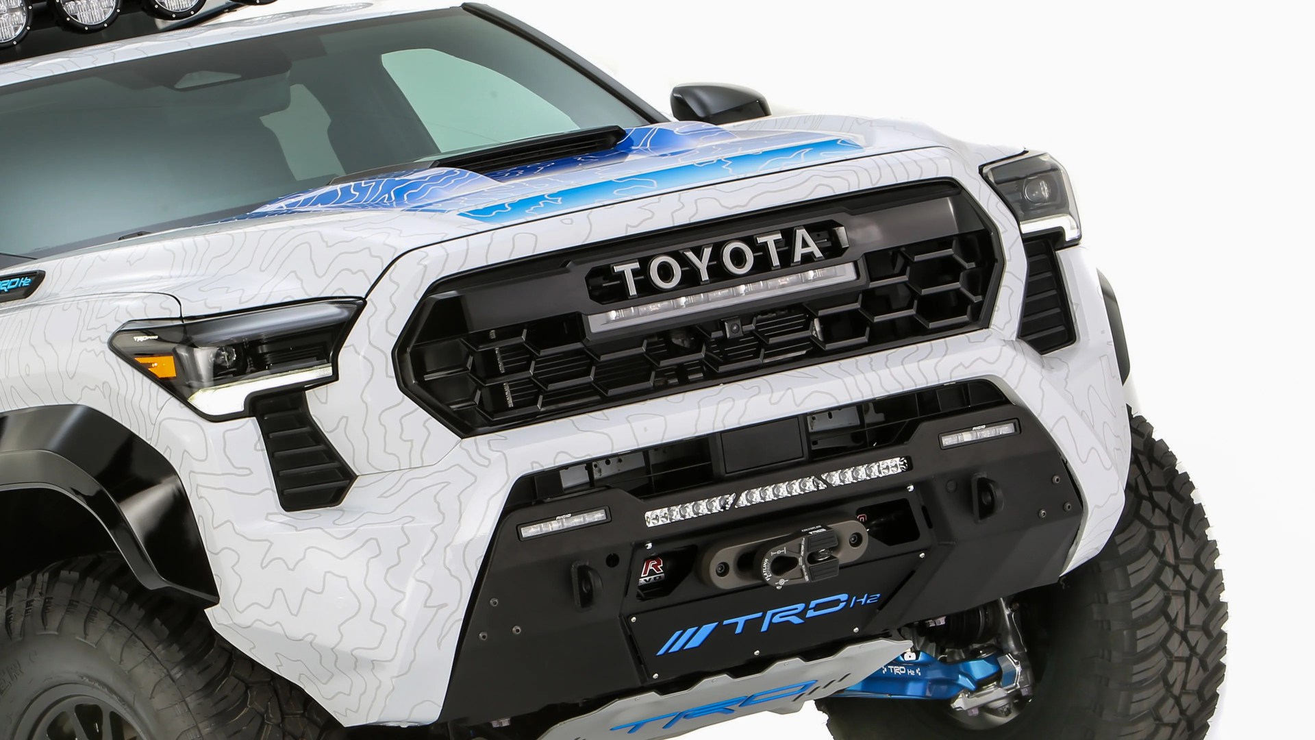 Front view of a white Toyota truck with black grille, blue hood accents, off-road tires, and TRD H2 branding on the bumper.