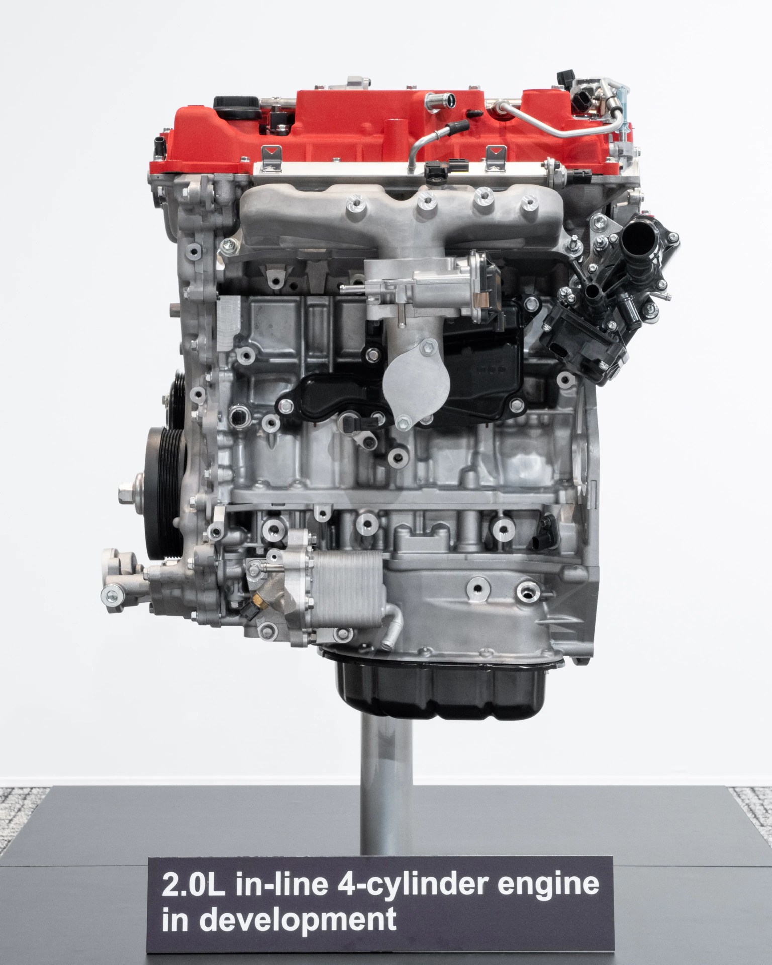 2.0L in-line 4-cylinder engine with red valve cover displayed on a stand with a sign.