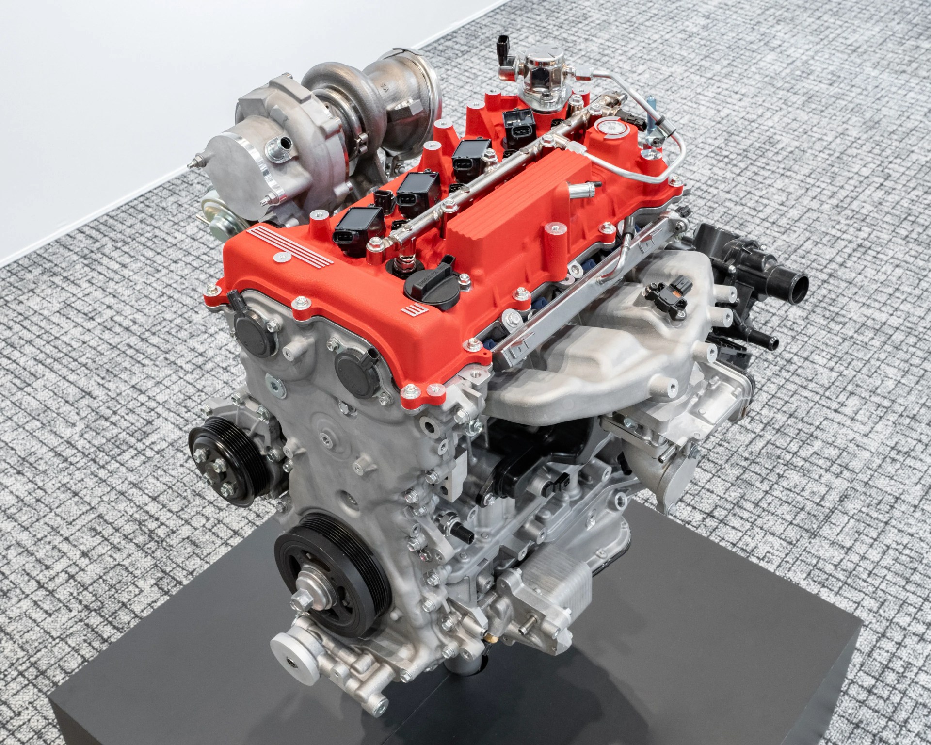 Silver car engine with a red valve cover and turbocharger mounted on a black platform.