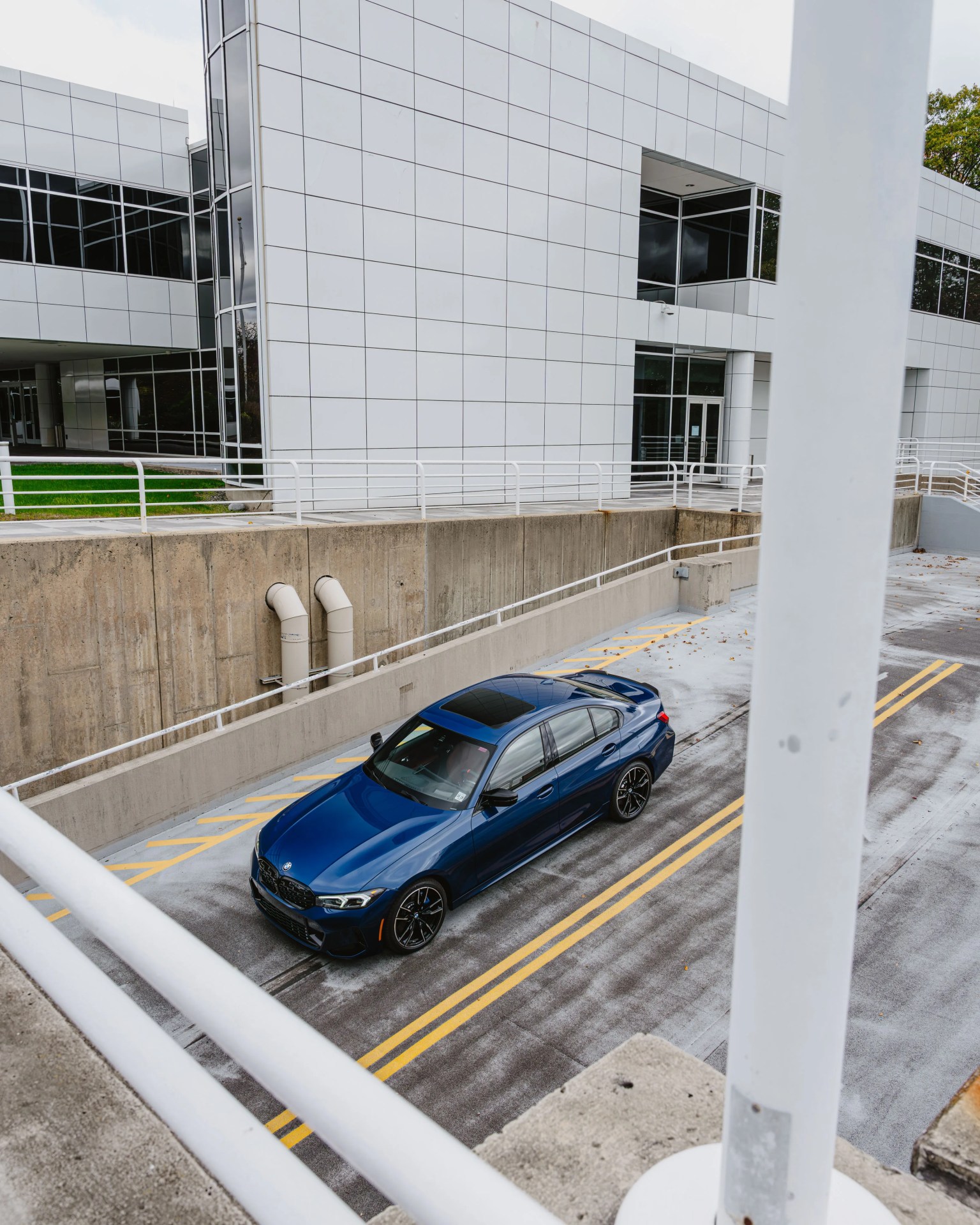 Blue BMW sedan parked in a concrete driveway next to a modern white building with large windows.