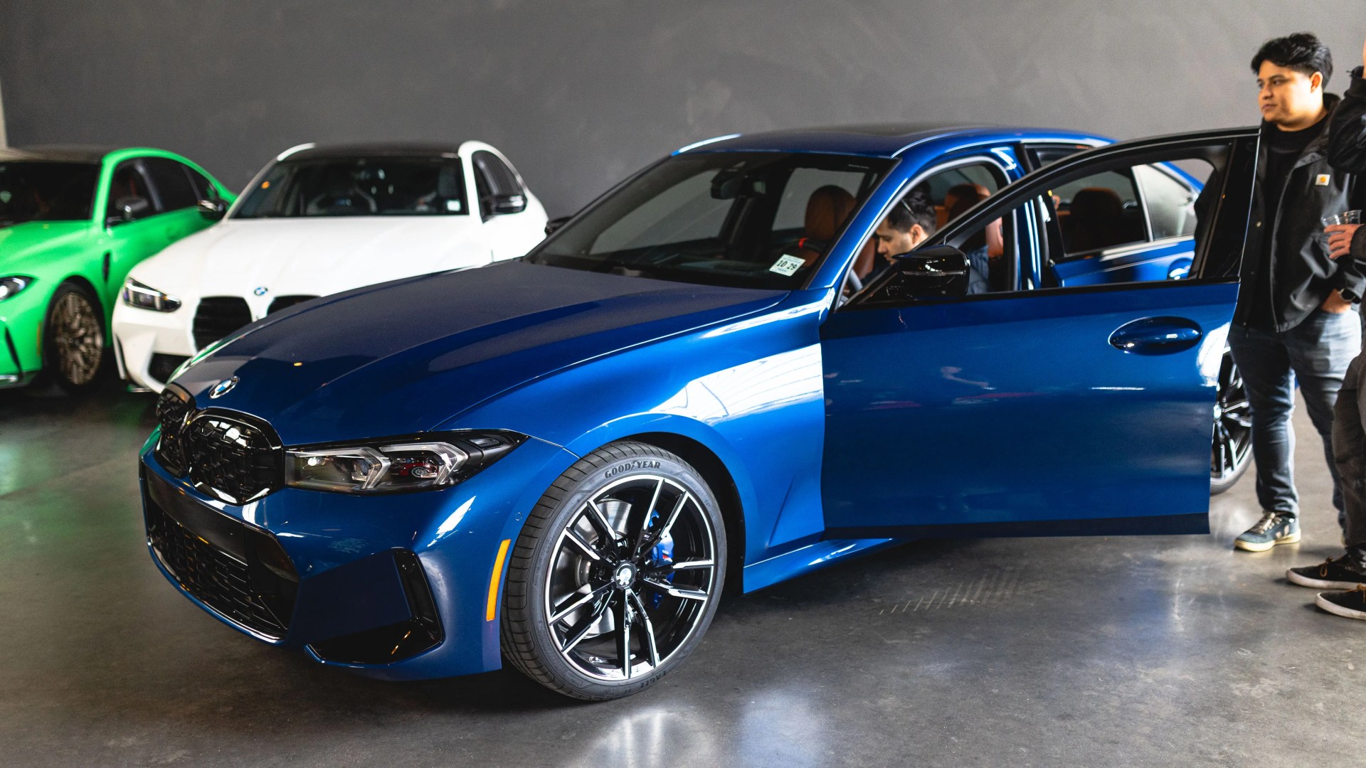 Blue BMW sedan with front passenger door open in an indoor showroom with other cars and people nearby.
