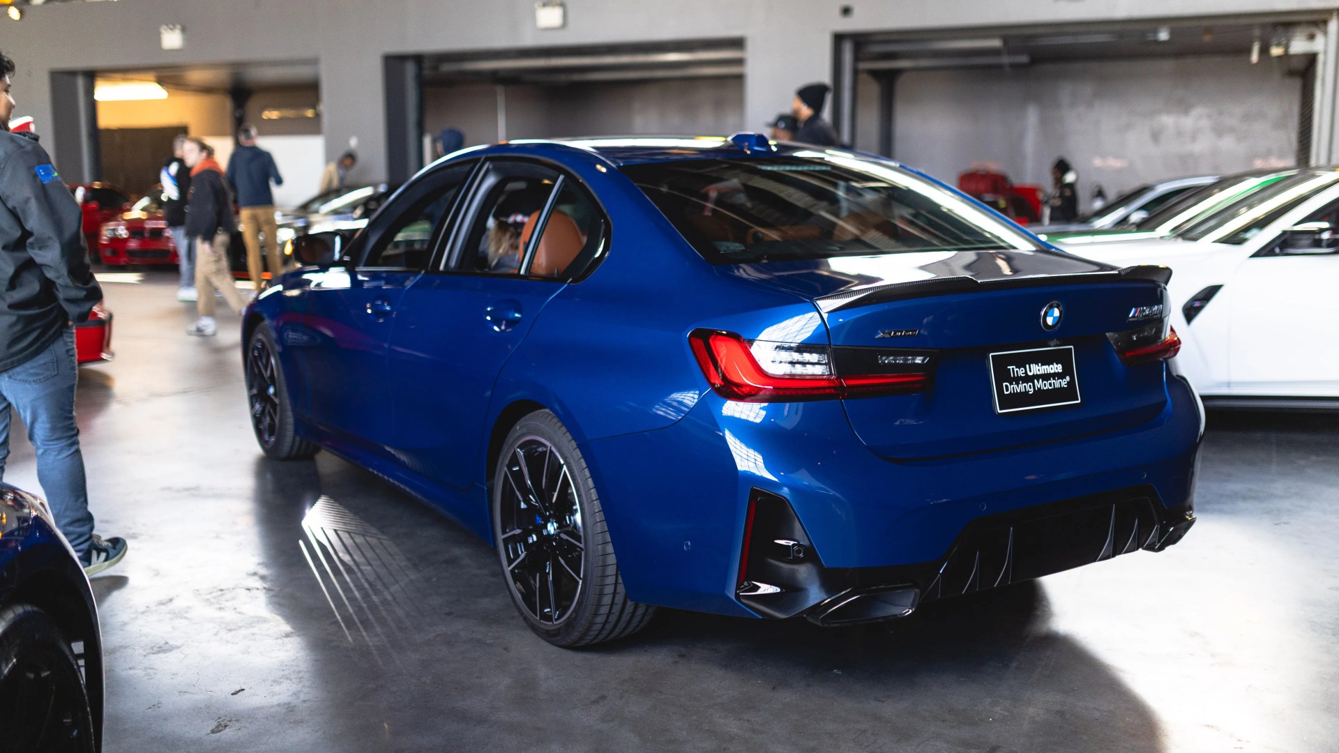Blue BMW M3 sedan with black wheels parked indoors among people and other cars.