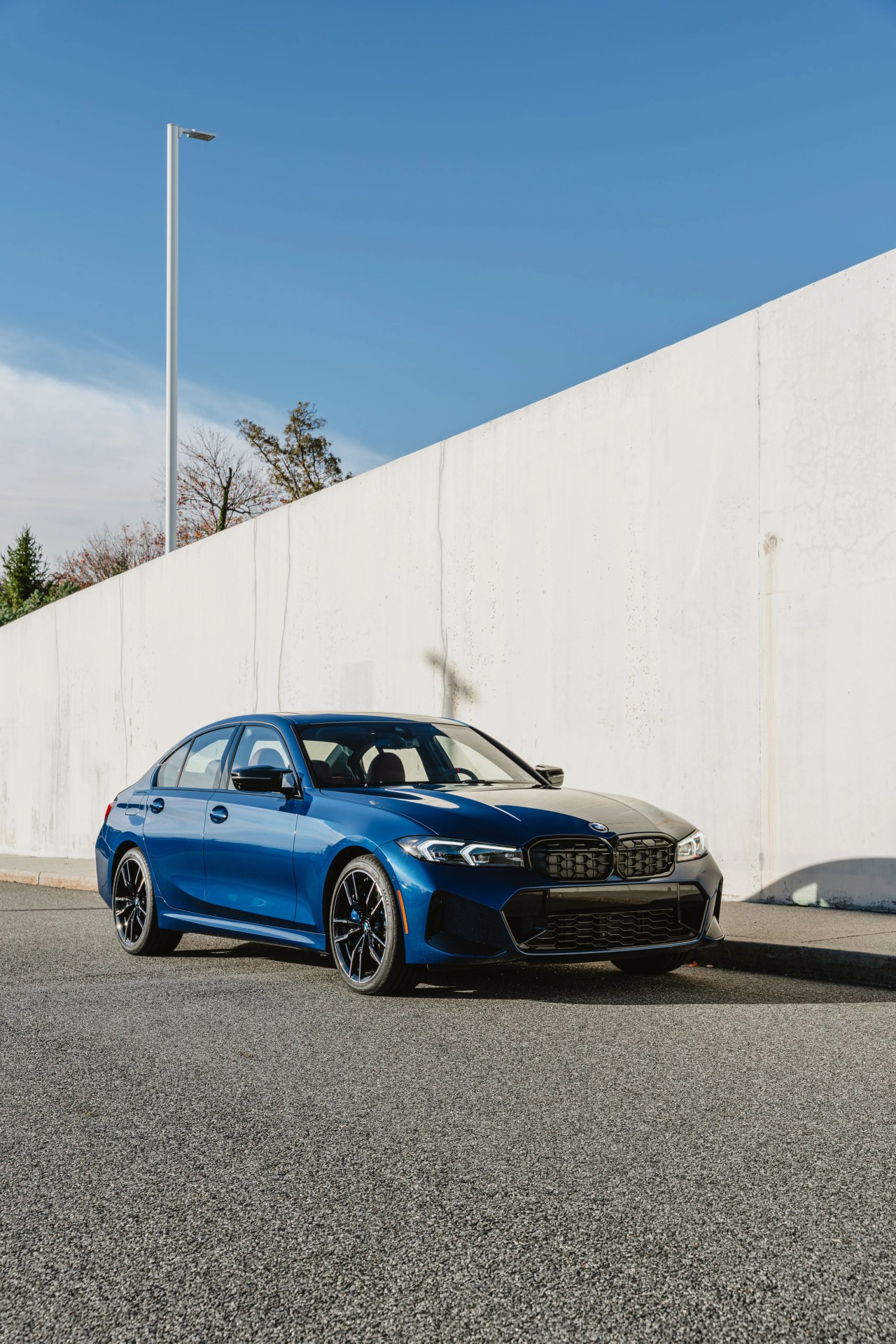 Blue BMW sedan with black grille and wheels parked on a street beside a white concrete wall.
