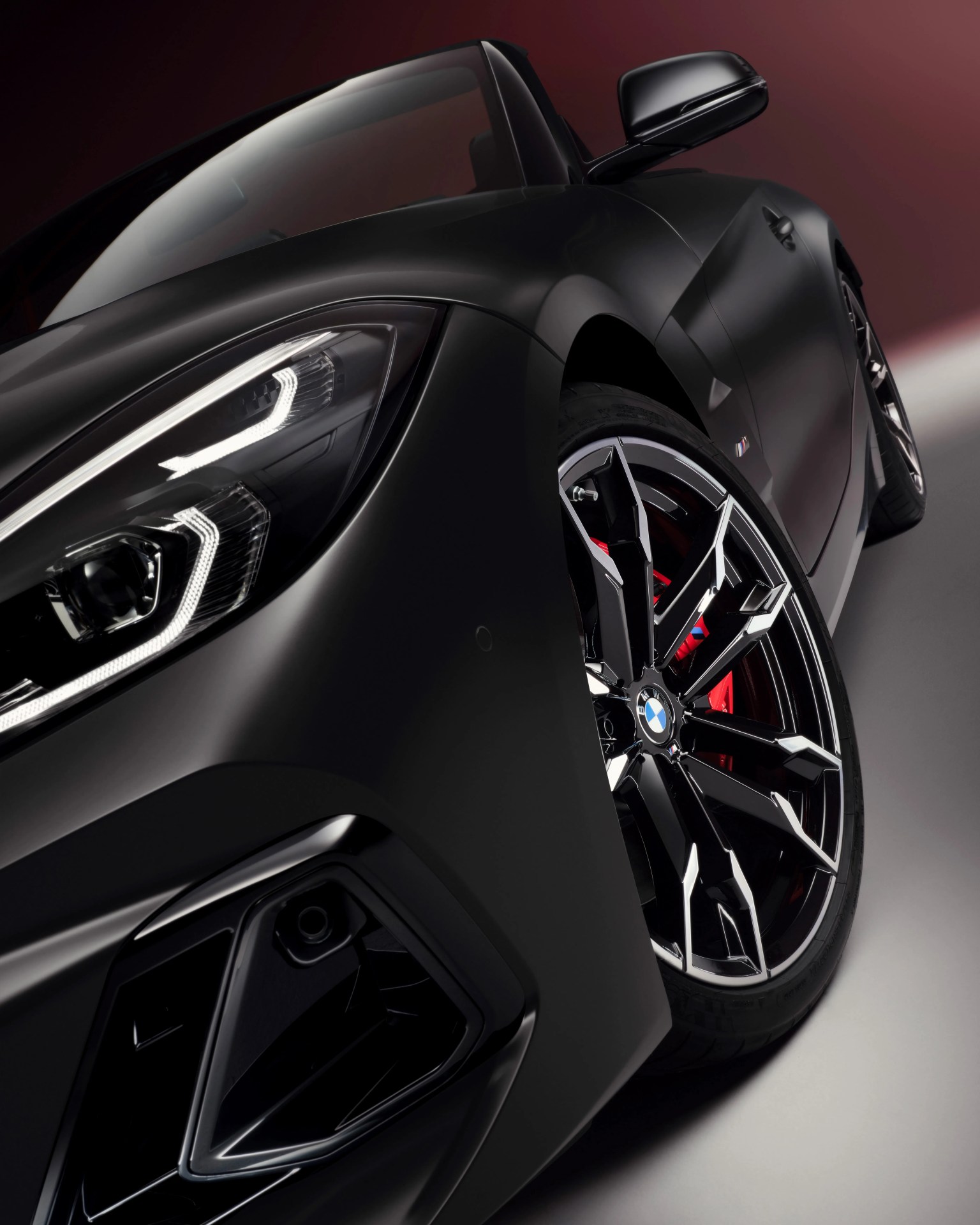 Close-up of a matte black BMW car front with detailed headlight and black alloy wheel with red brake caliper.