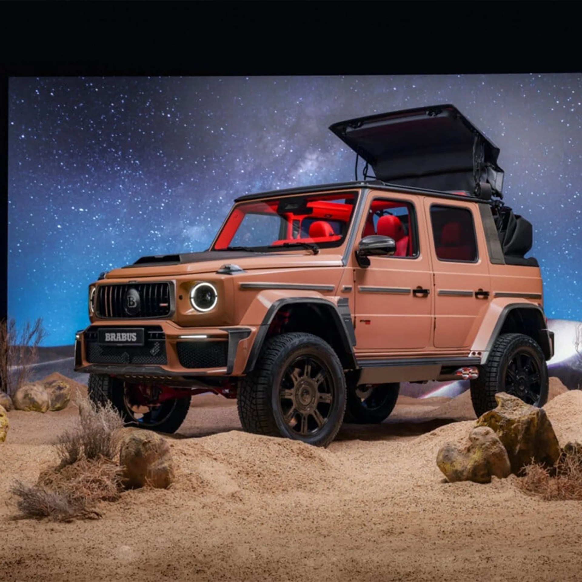 Brabus-modified tan off-road SUV with red interior and open roof tent on sandy terrain under starry sky.