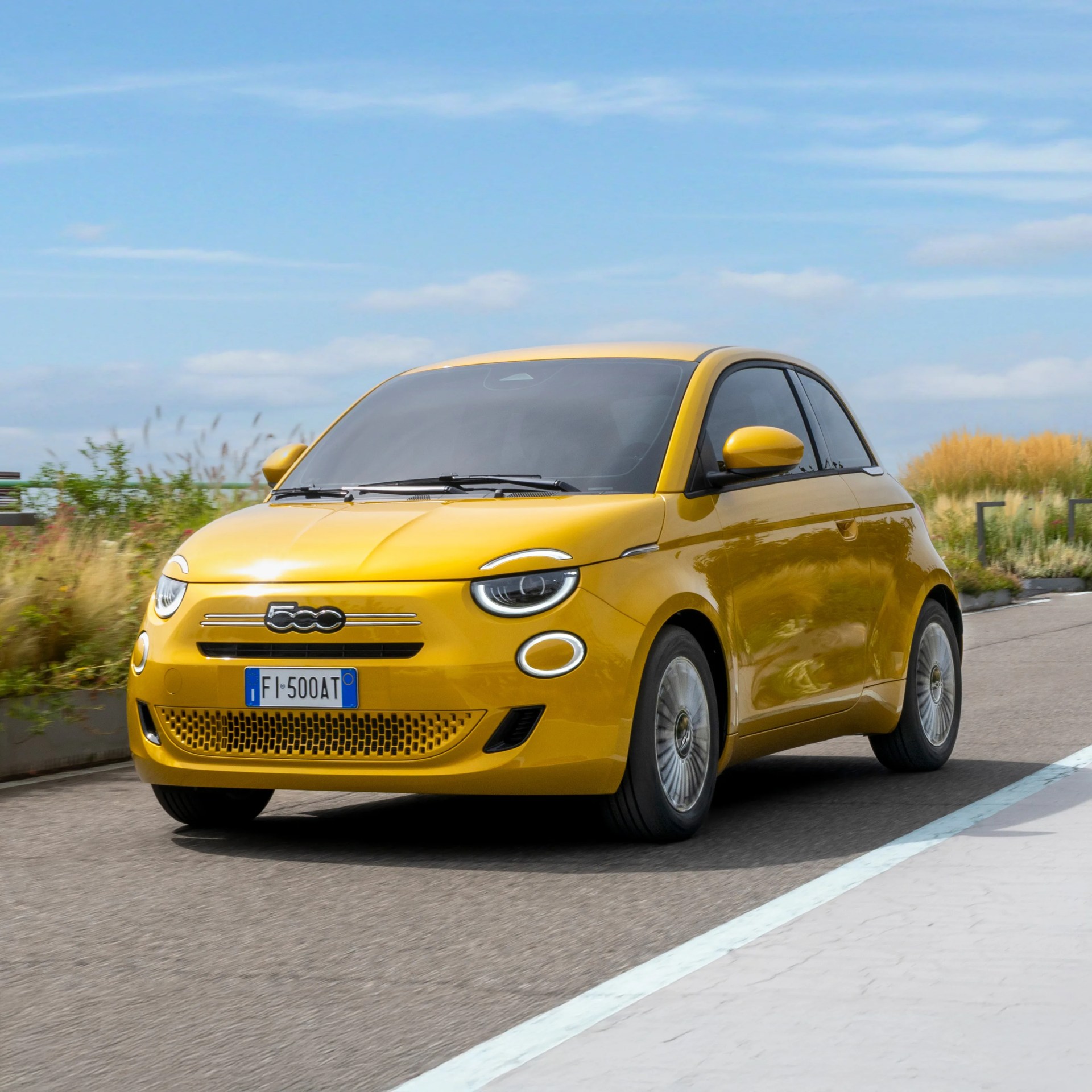 Yellow Fiat 500 compact car parked on a road with greenery and blue sky in the background.