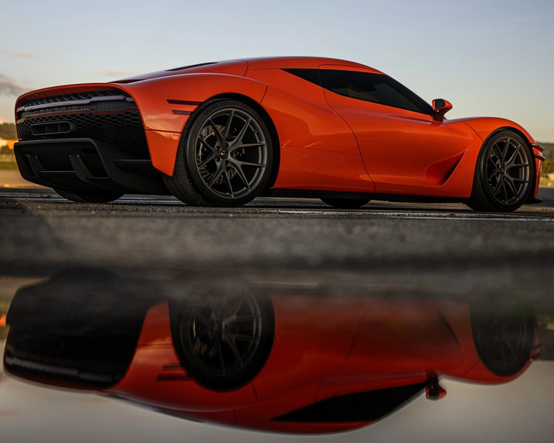 Orange sports car with black wheels parked on asphalt, reflected in a puddle below.