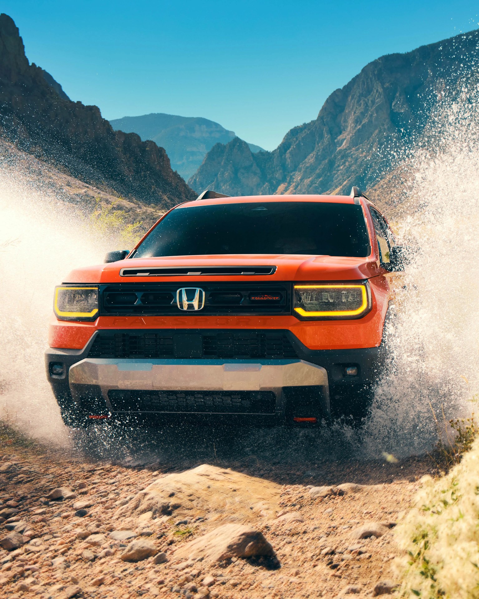 Orange Honda SUV splashing through water on a rocky mountain trail under clear blue sky.