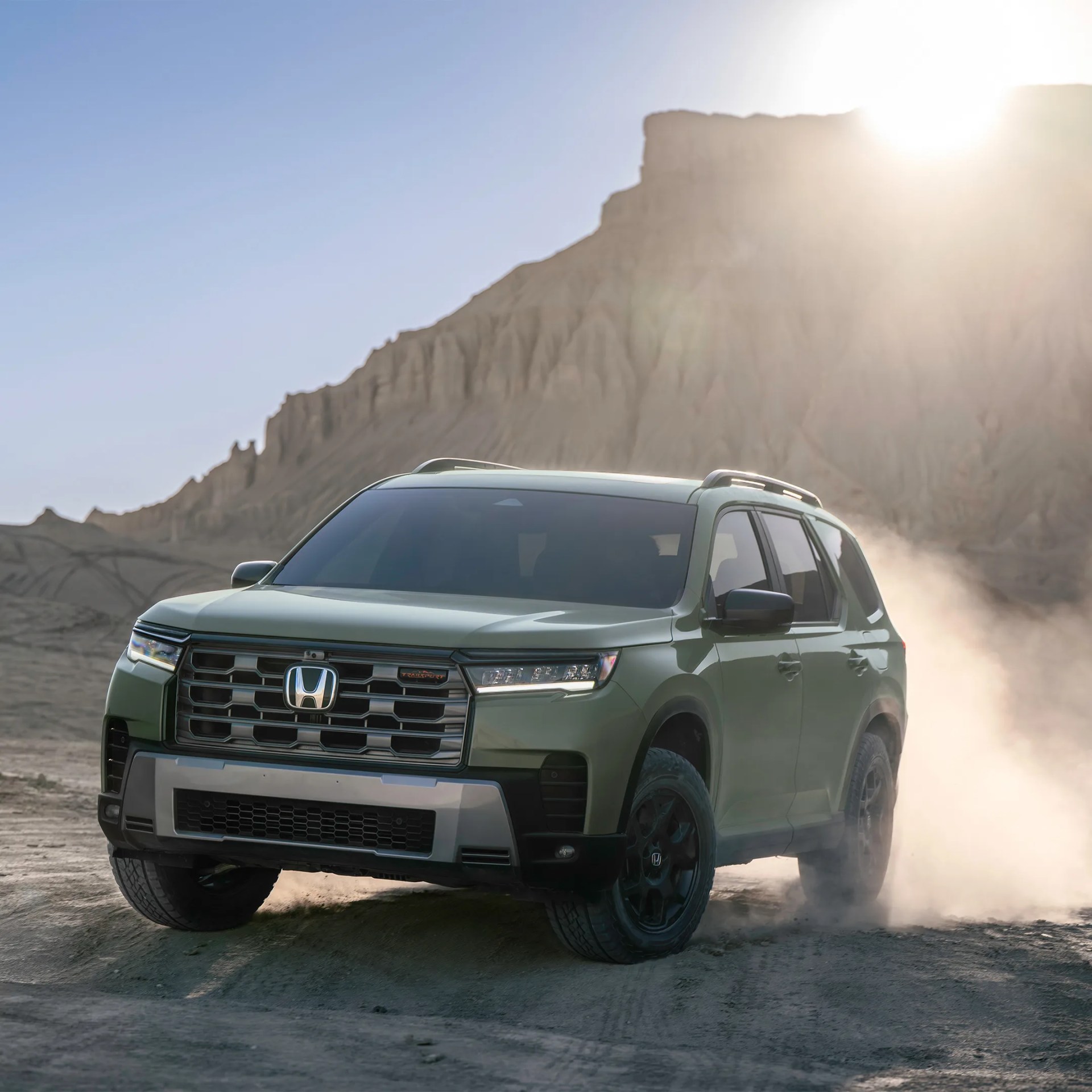 Green Honda SUV with black wheels kicking up dust in a desert landscape at sunset.
