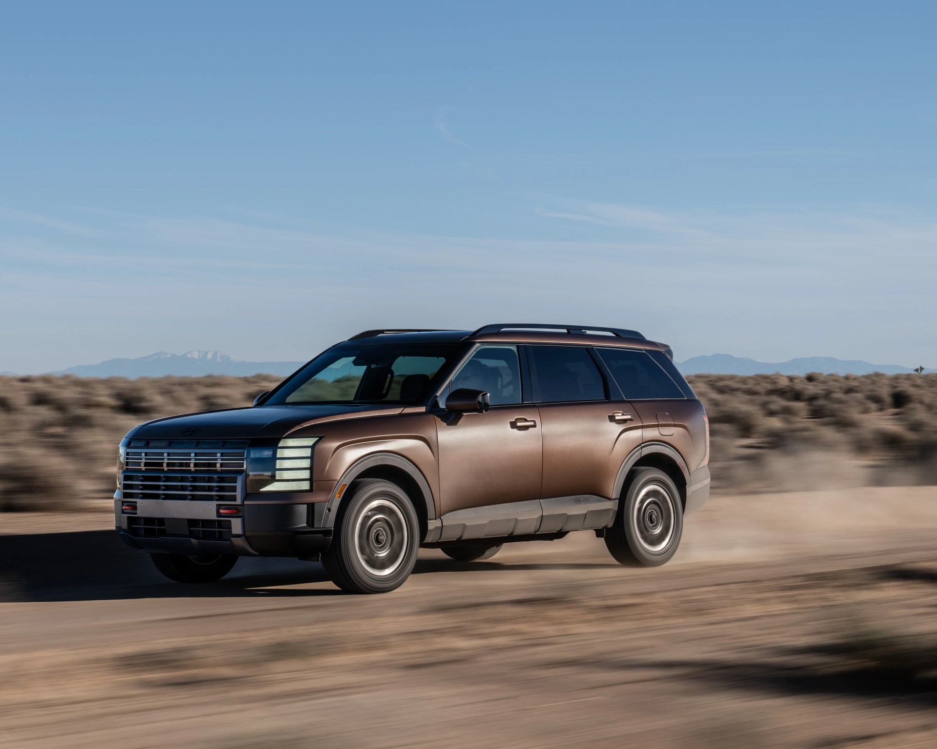 Brown electric SUV driving on a dusty desert road with mountains in the background.