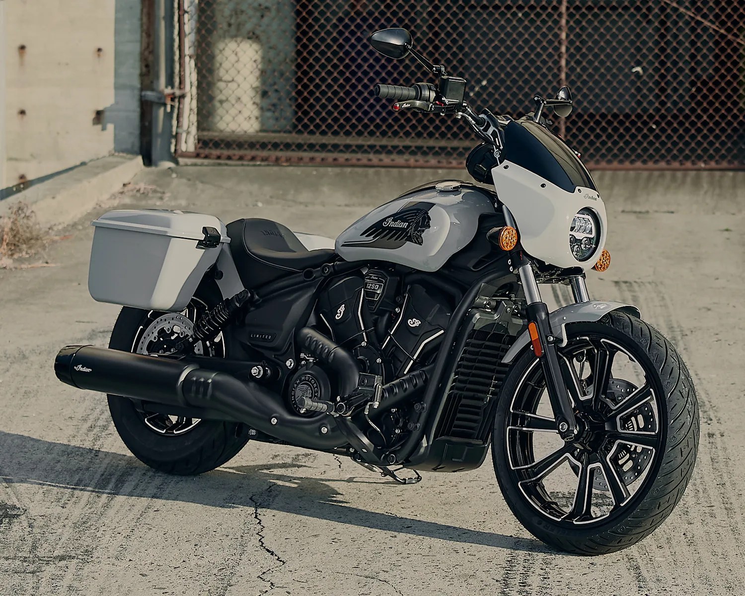 Gray and black Indian motorcycle with saddlebags parked on concrete near a chain-link fence.
