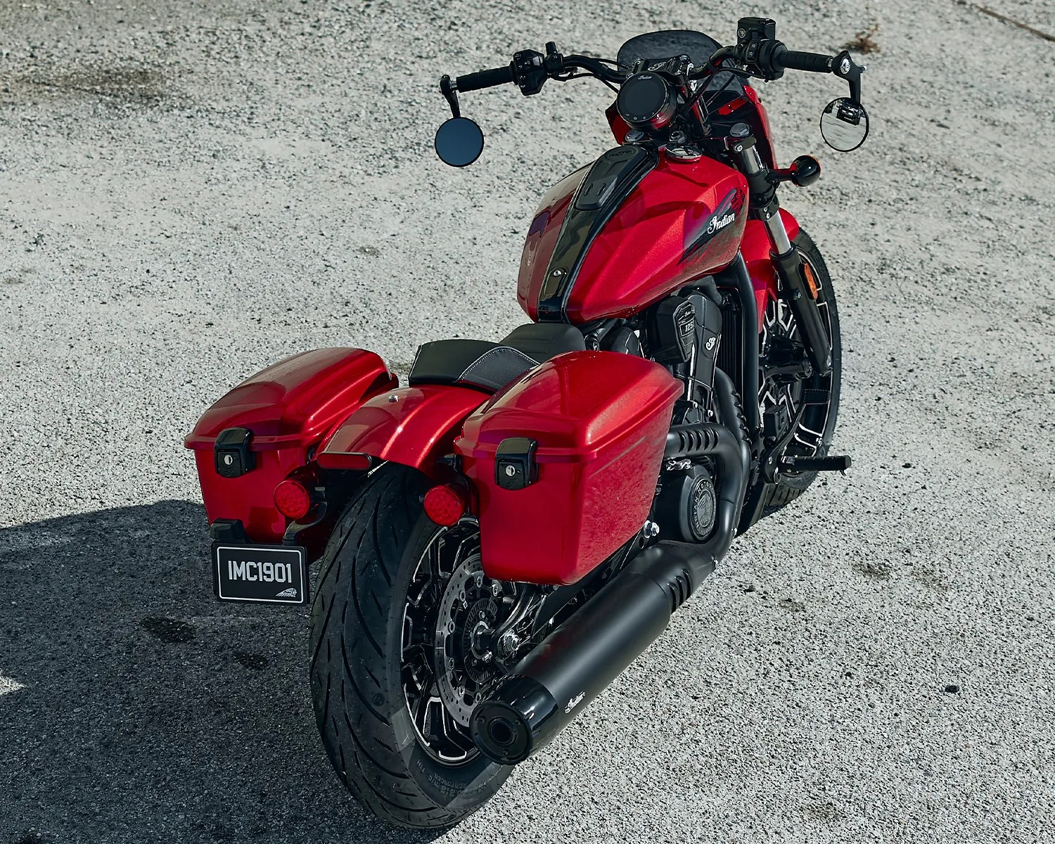 Red Indian motorcycle with black exhaust pipes and saddlebags parked on a gravel surface.