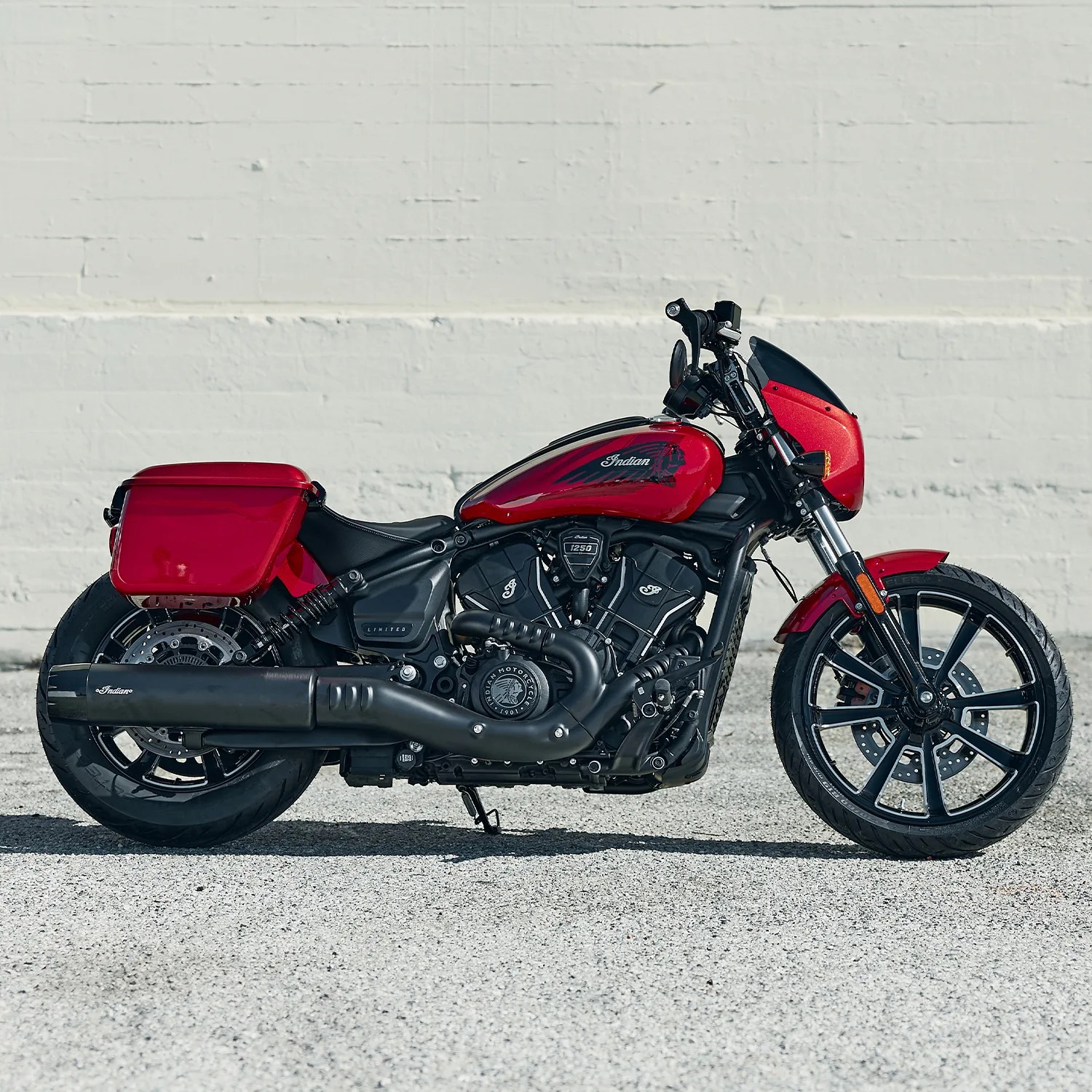 Red Indian motorcycle with black engine, exhaust, and wheels parked on gravel in front of a white brick wall.