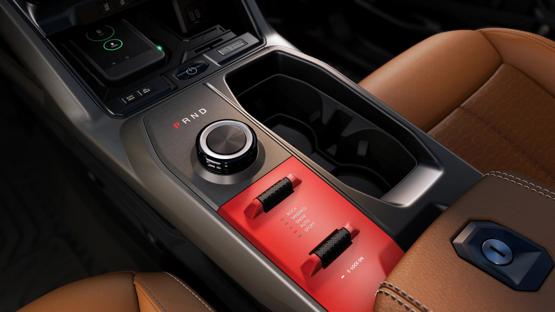 Car center console with rotary gear selector, dual cup holders, red terrain mode panel, and brown leather seats.