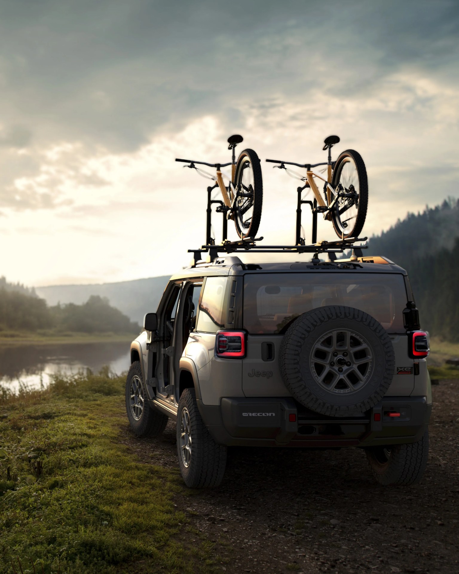 Gray Jeep SUV with two mountain bikes mounted on the roof rack parked on a dirt path near a lake at sunset.