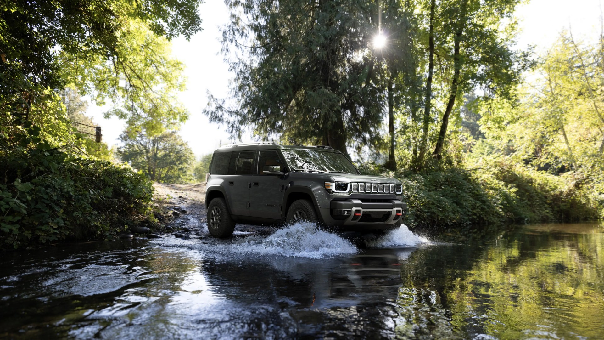 Dark gray Jeep SUV driving through shallow water in a forested area with sunlight filtering through trees.