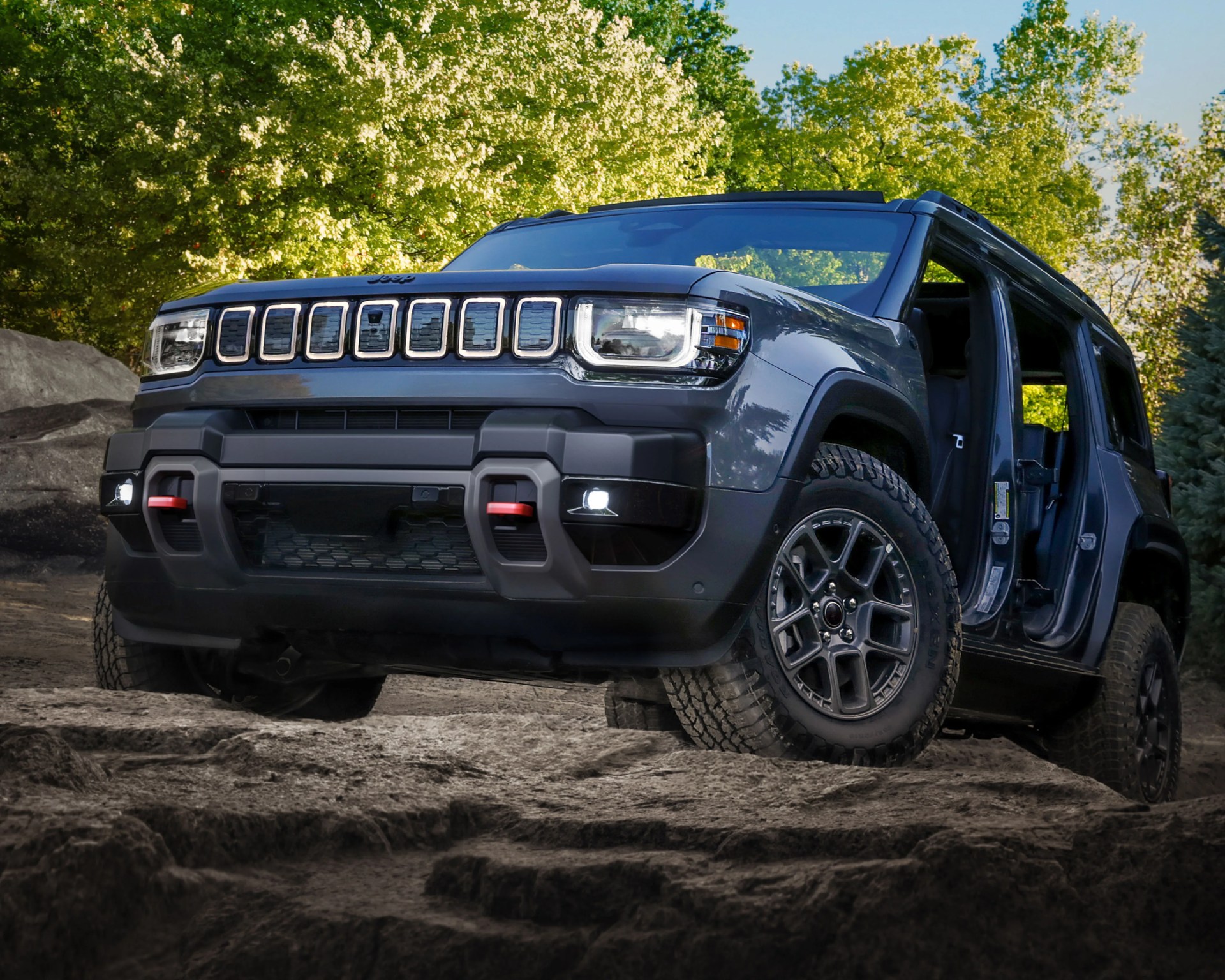 Dark gray Jeep SUV with off-road tires and open doors on rocky terrain with green trees in the background.