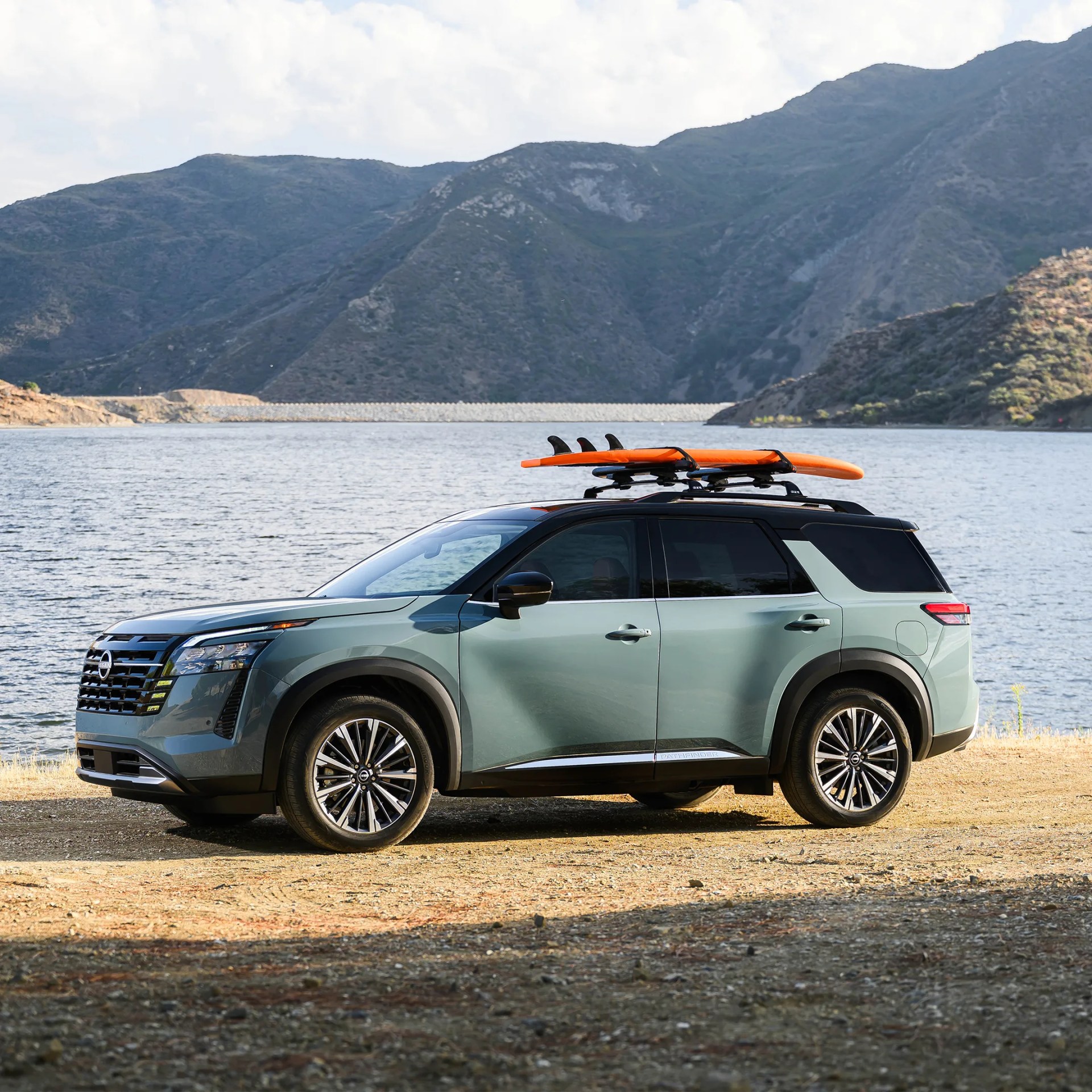 Green Nissan SUV with black roof and orange surfboards mounted on roof racks, parked by a lake with mountains in the background.