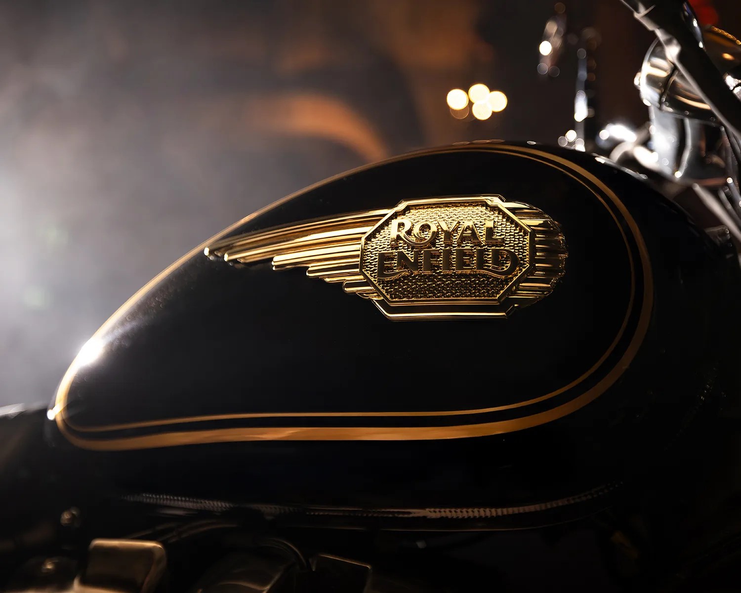 Glossy black motorcycle fuel tank with gold Royal Enfield emblem and gold pinstriping.