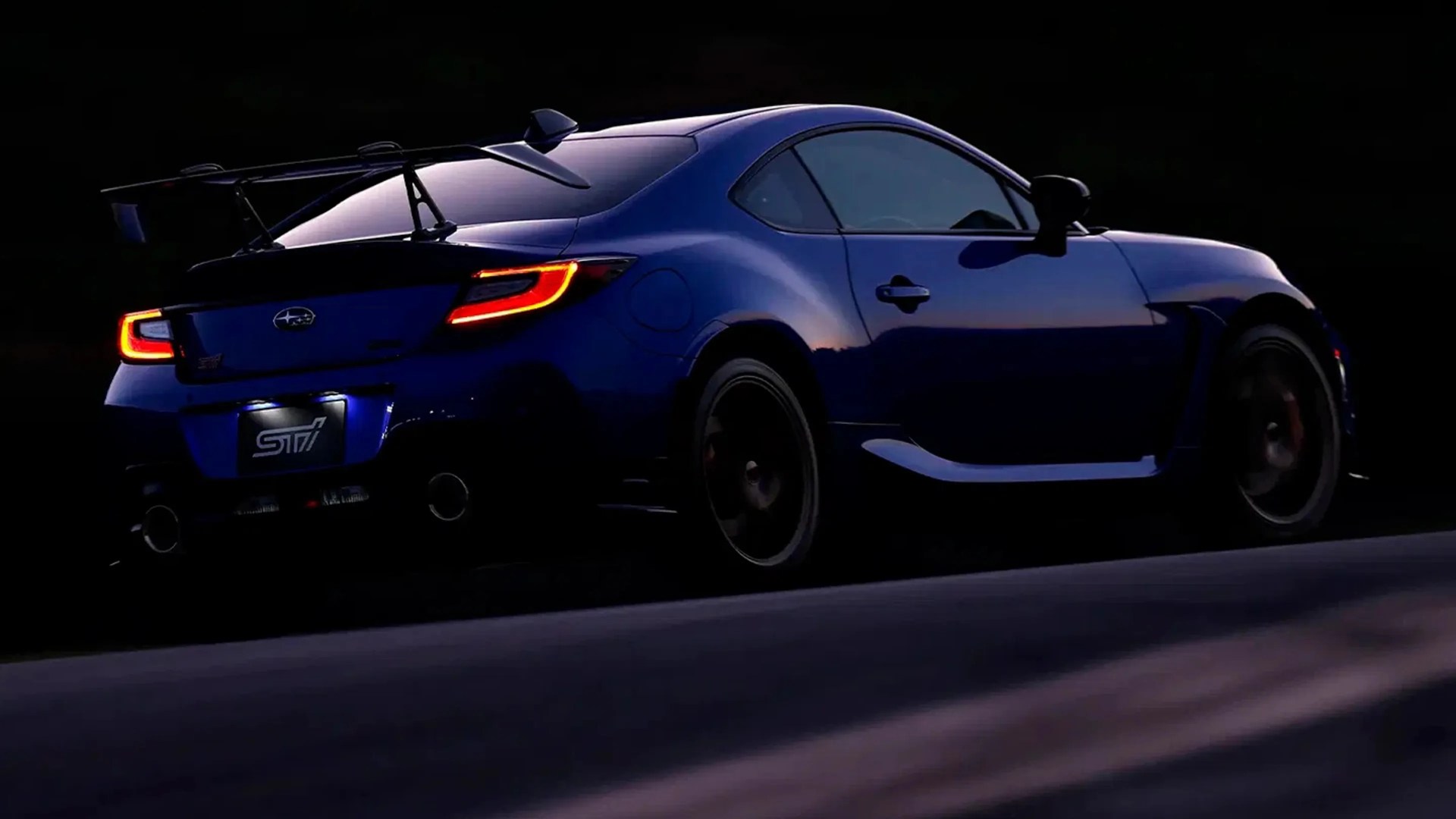 Blue Subaru sports car with illuminated taillights and large rear spoiler at dusk.