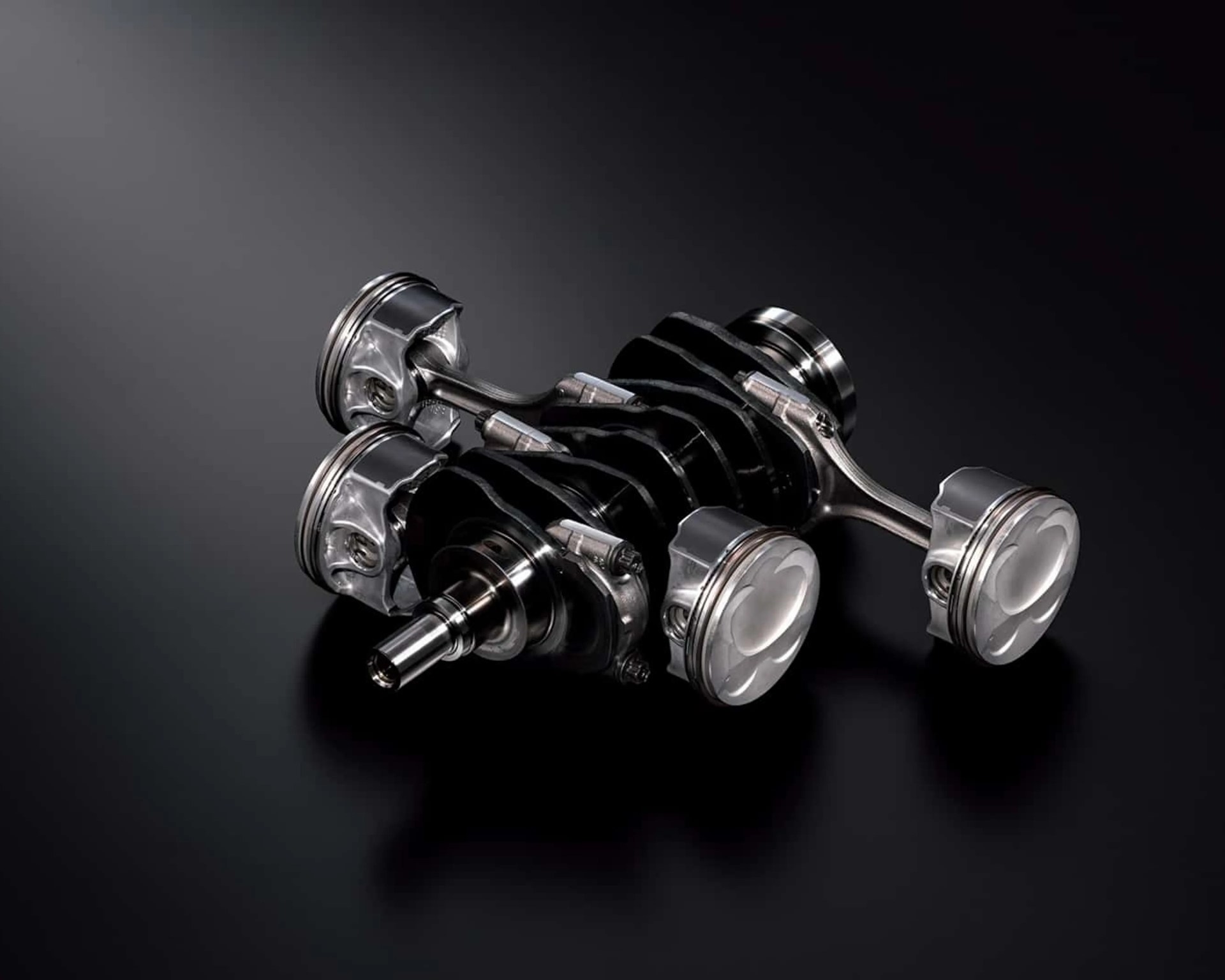 Metal crankshaft with four attached pistons on a dark reflective surface.