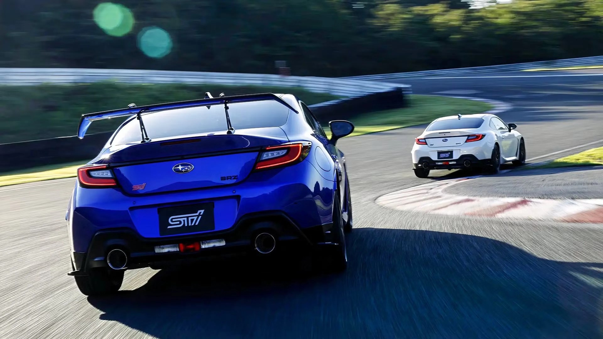 Blue Subaru BRZ STI with large rear wing and white Subaru BRZ STI on a racetrack corner.