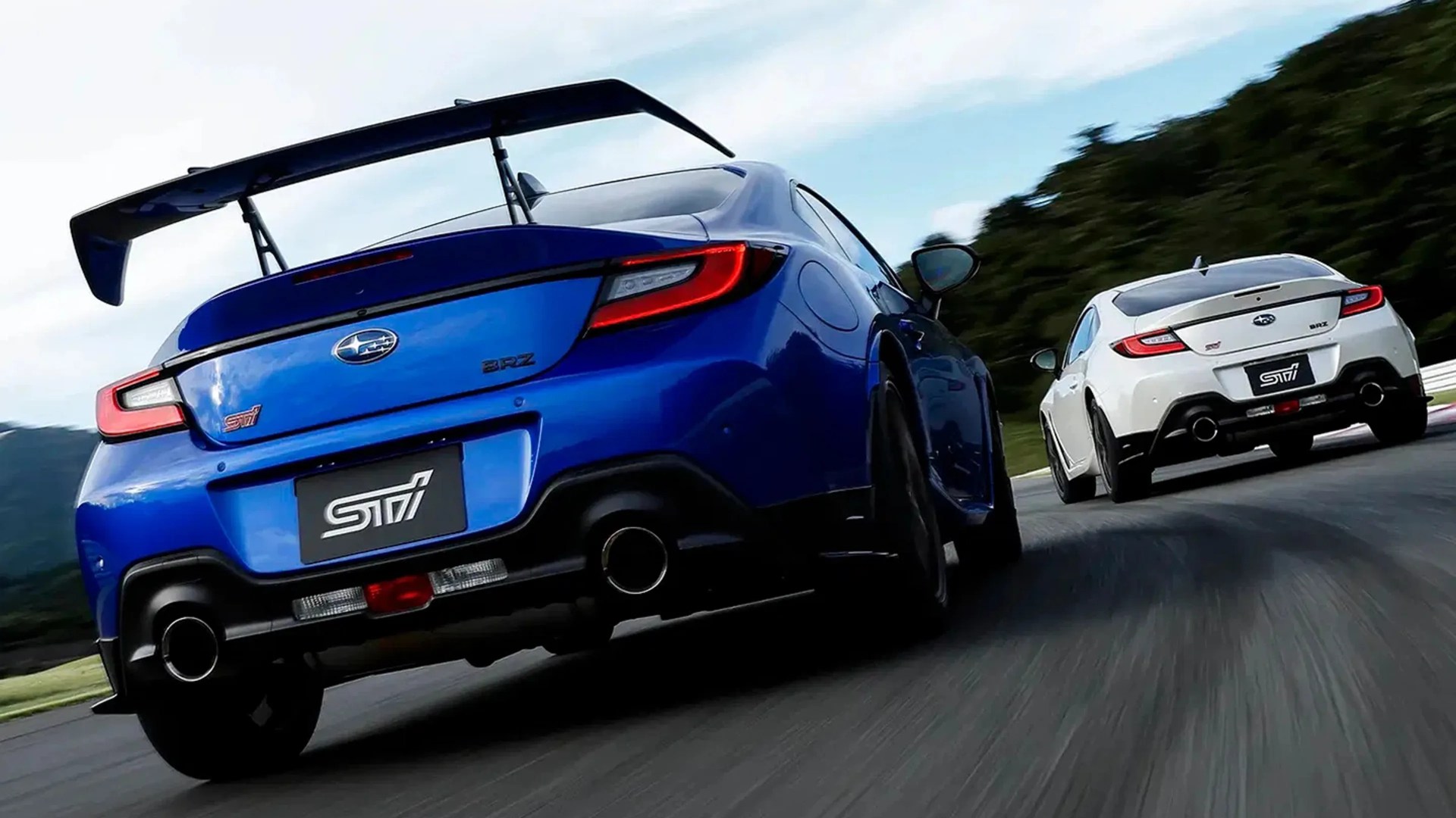 Two Subaru BRZ STI sports cars, one blue with a large rear wing and one white, driving on a racetrack.