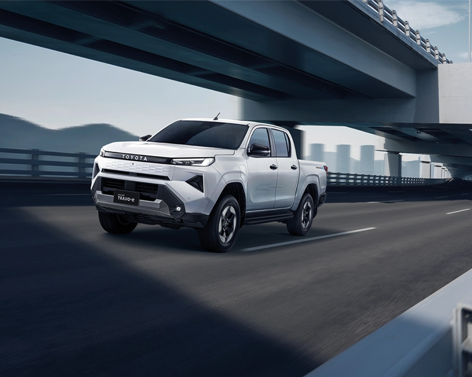 White Toyota Hilux TRAVO-e pickup truck driving on a highway under a bridge.