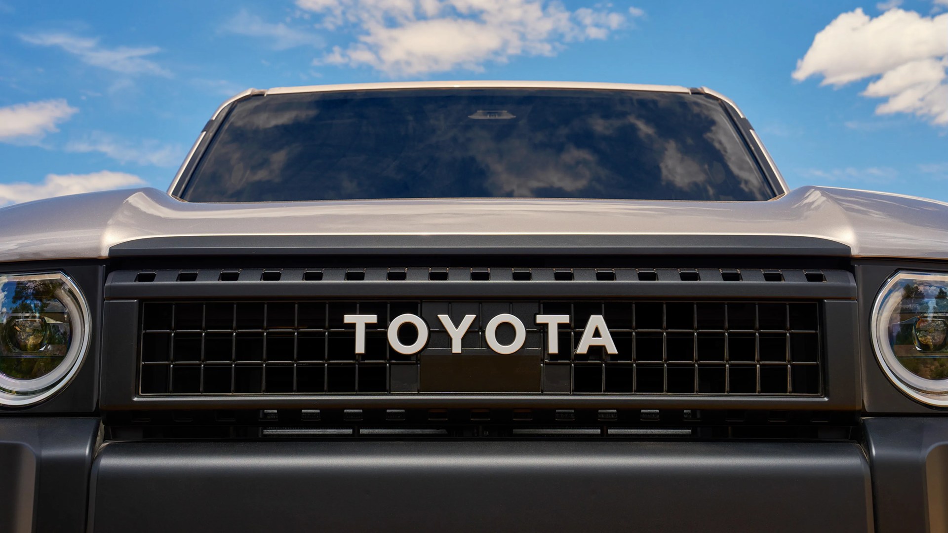 Front grille of a silver Toyota vehicle with bold white TOYOTA lettering and round headlights under a blue sky.