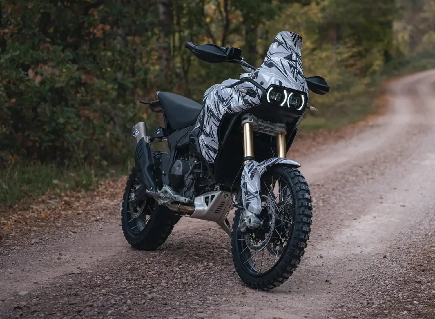 Adventure motorcycle with black and gray geometric camouflage pattern parked on a dirt road surrounded by trees.