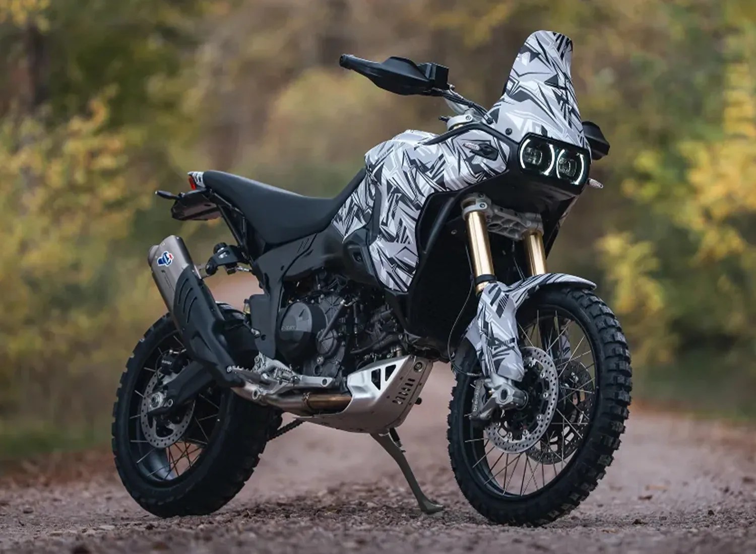 Dual-sport motorcycle with black and white geometric camouflage pattern parked on a dirt road.
