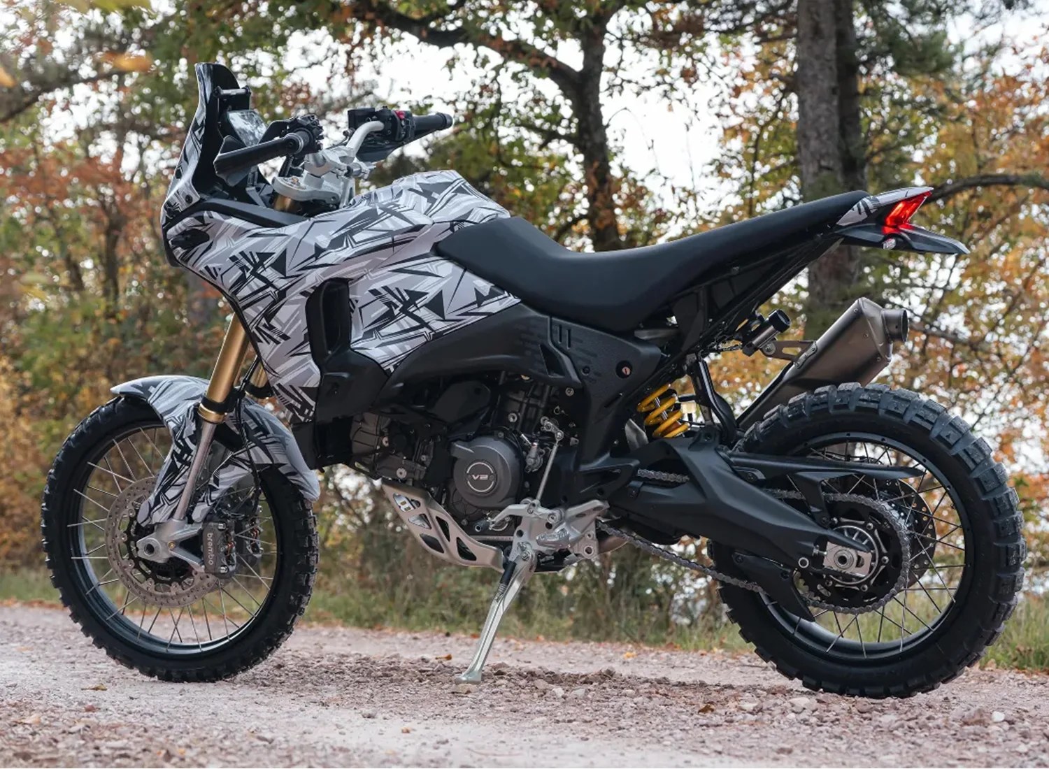 Adventure motorcycle with black and white geometric camo pattern, knobby tires, and gold front forks on a dirt path.