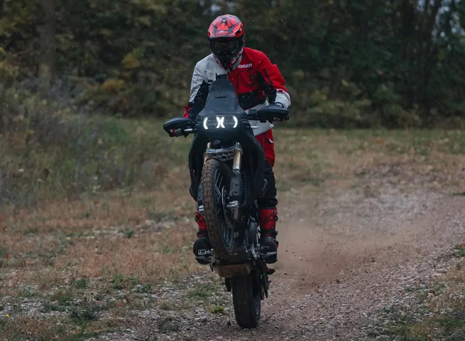 Motorcyclist in red and white gear performing a wheelie on a black off-road motorcycle on a dirt path.