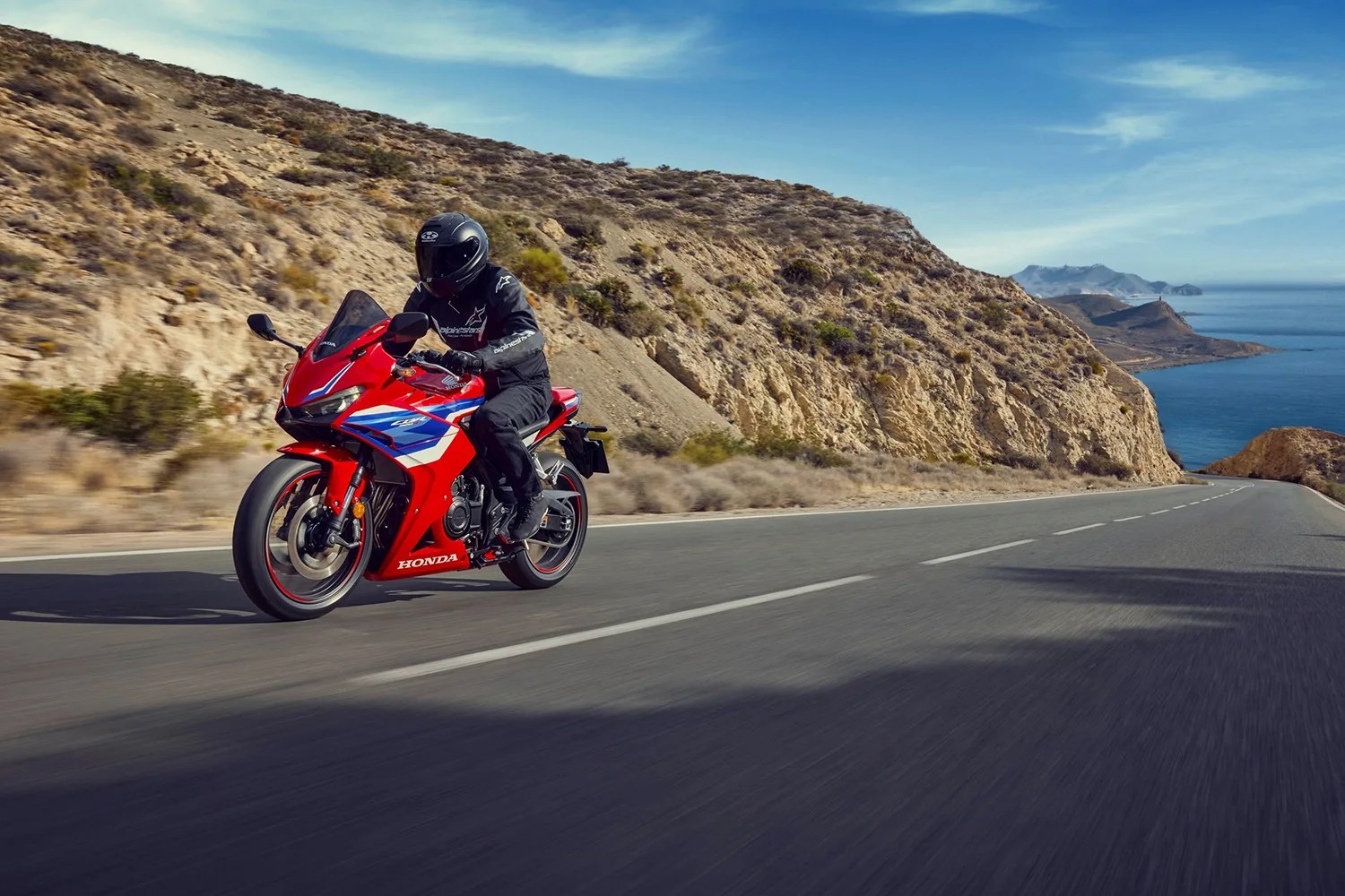 Red Honda sport motorcycle with blue and white accents ridden on a coastal road with rocky hills and ocean in the background.