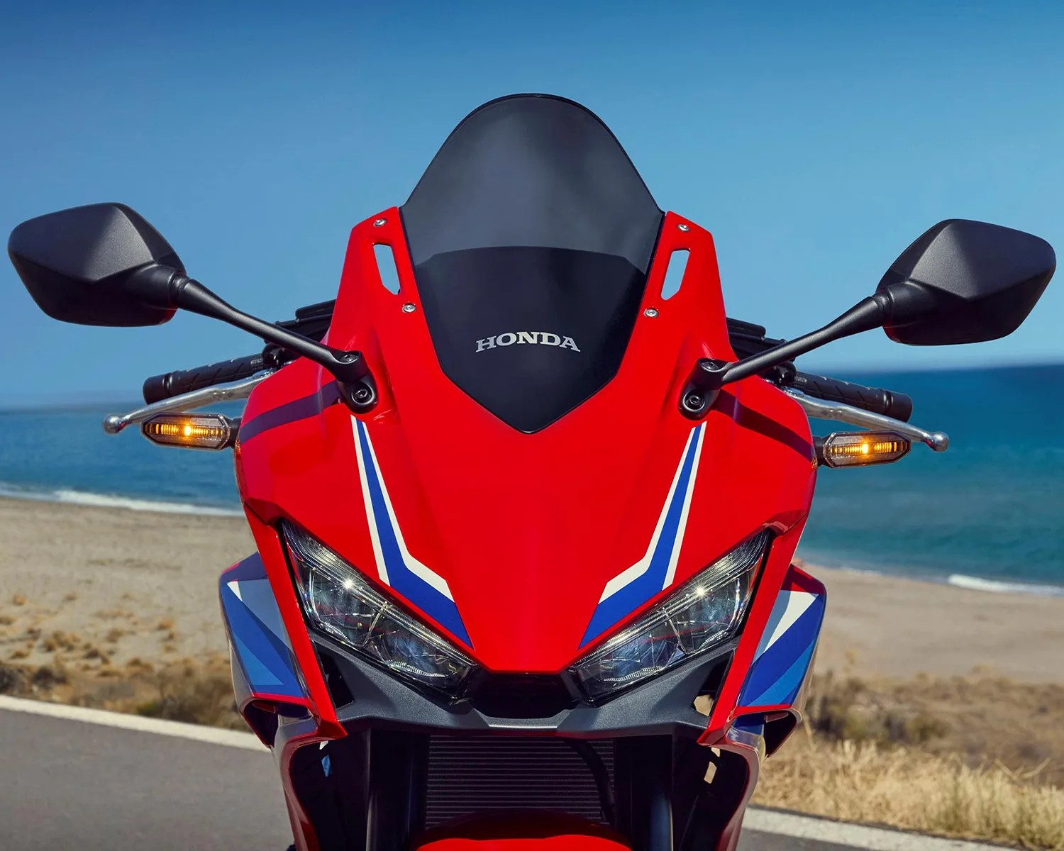 Front view of a red Honda sport motorcycle with blue and white stripes, black mirrors, and turn signals on a coastal road.