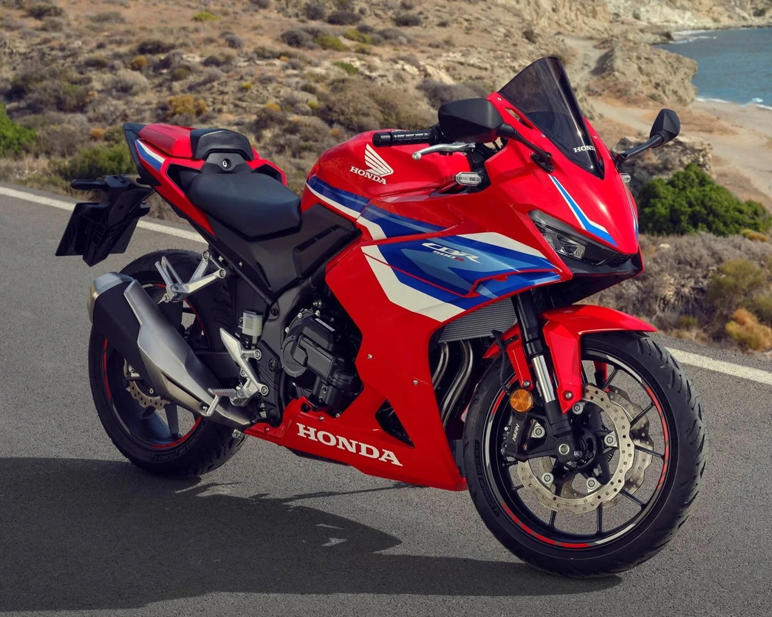 Red Honda CBR sport motorcycle with blue and white accents parked on a road near a rocky coastline.