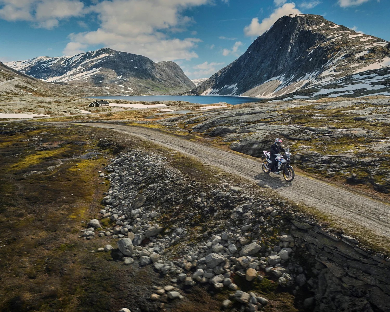 Motorcyclist riding a dual-sport bike on a gravel road in a mountainous landscape with patches of snow and a lake.