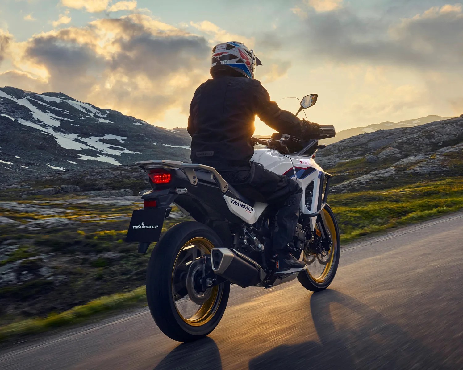 Person riding a white Honda Transalp 750 motorcycle with gold rims on a mountain road at sunset.