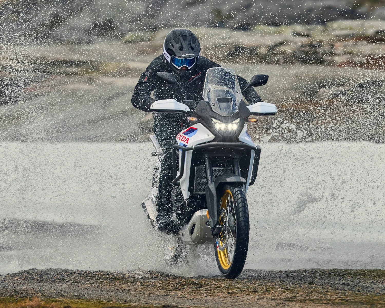 Motorcyclist wearing black gear riding a white Honda adventure bike through splashing water.