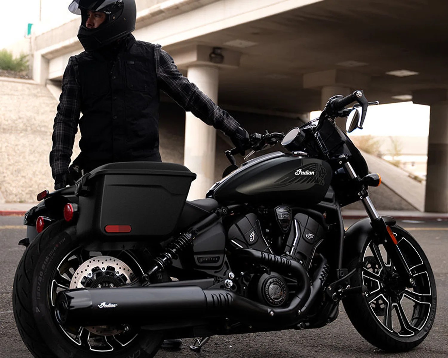 Black Indian motorcycle with matte finish and hard saddlebags parked under a bridge with a rider in black gear and helmet.