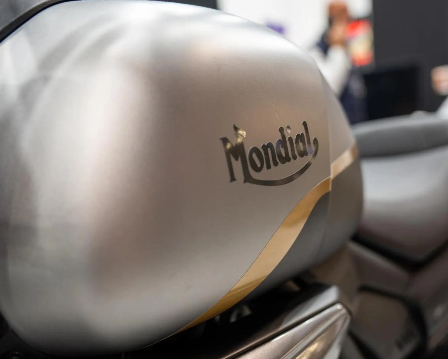 Close-up of a silver motorcycle fuel tank with the black "Mondial" logo and a gold stripe.