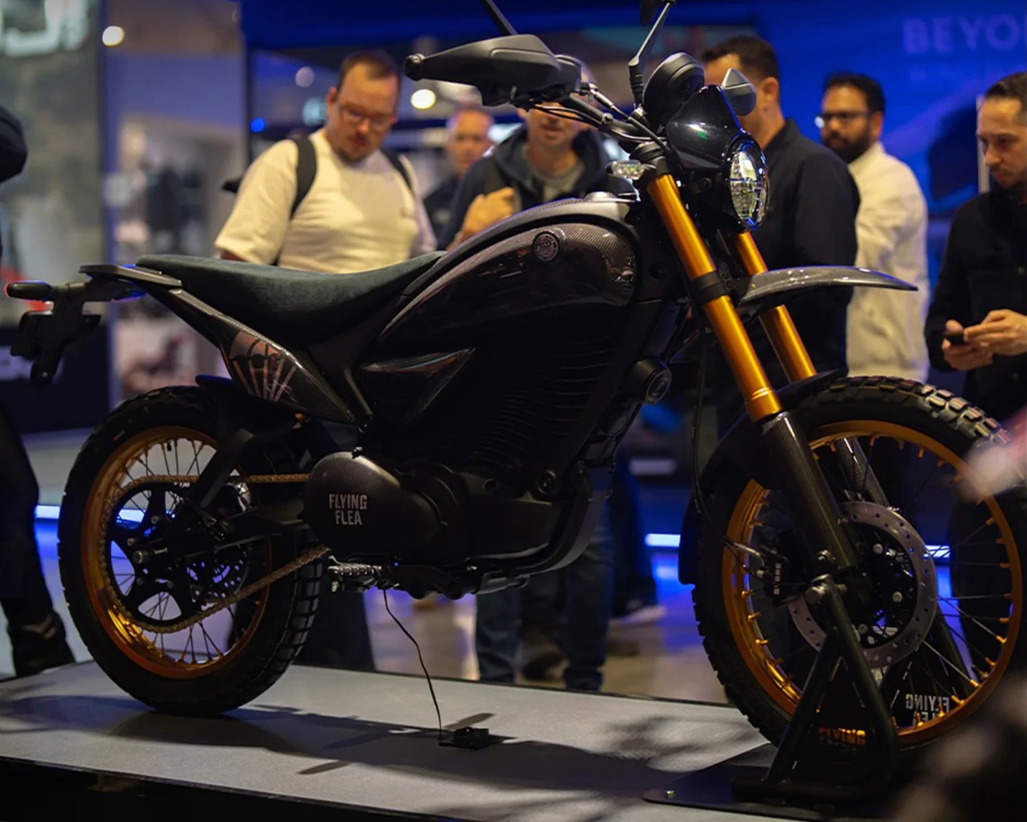 Black and gold Flying Flea motorcycle with knobby tires displayed indoors with people in the background.