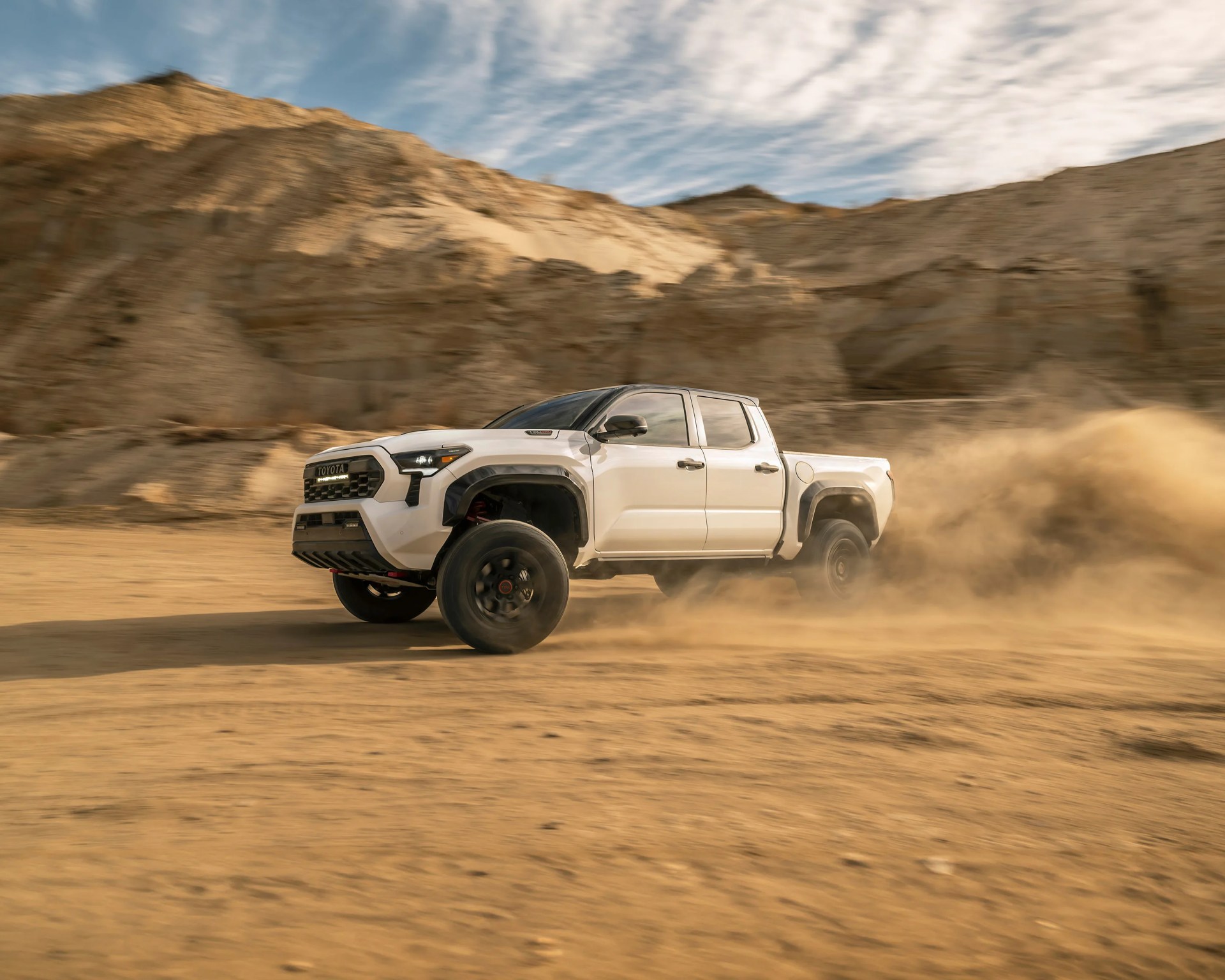 White Toyota pickup truck kicking up dust while driving fast on a desert terrain with rocky hills in the background.