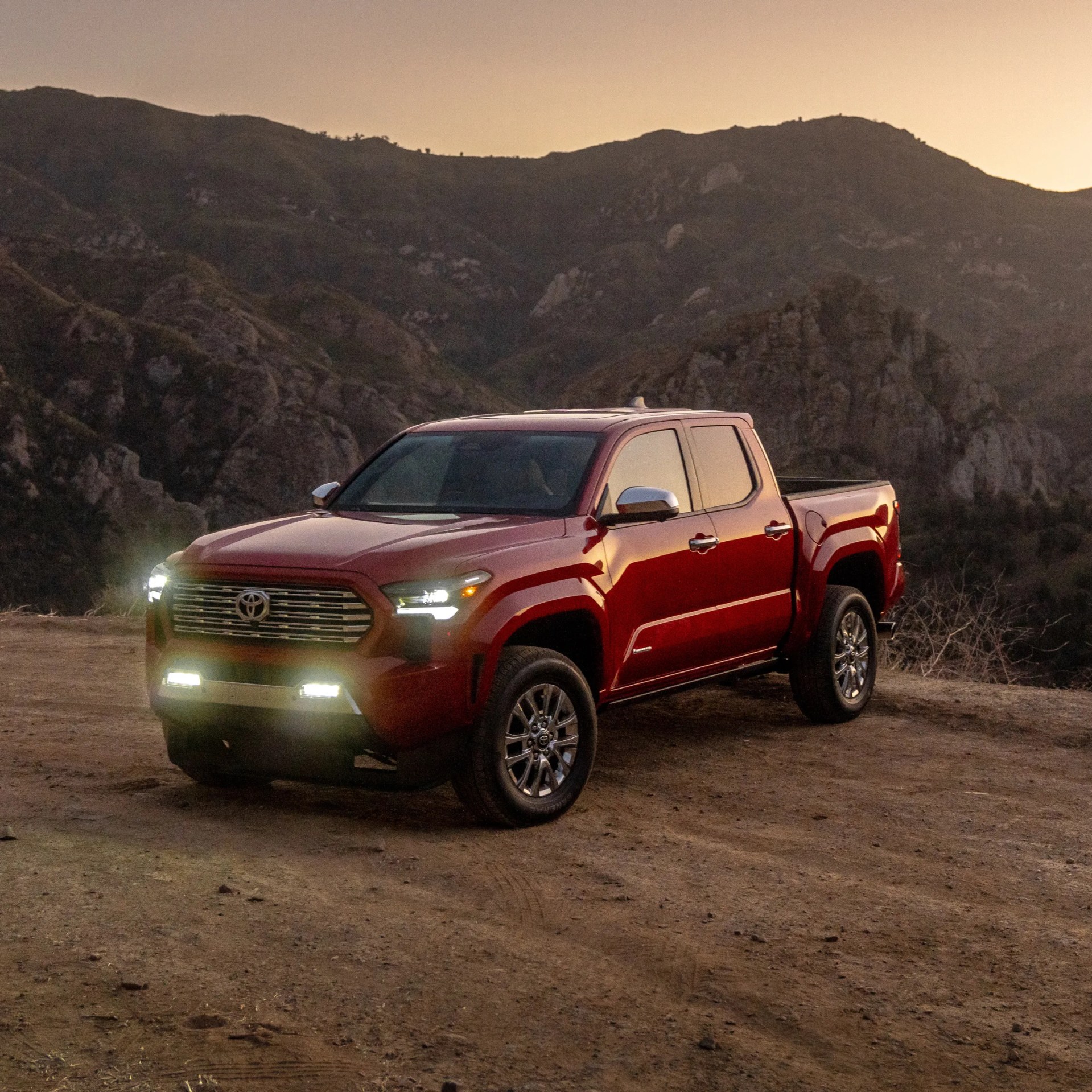 Red Toyota pickup truck with headlights on parked on a dirt road with rocky hills in the background at dusk.