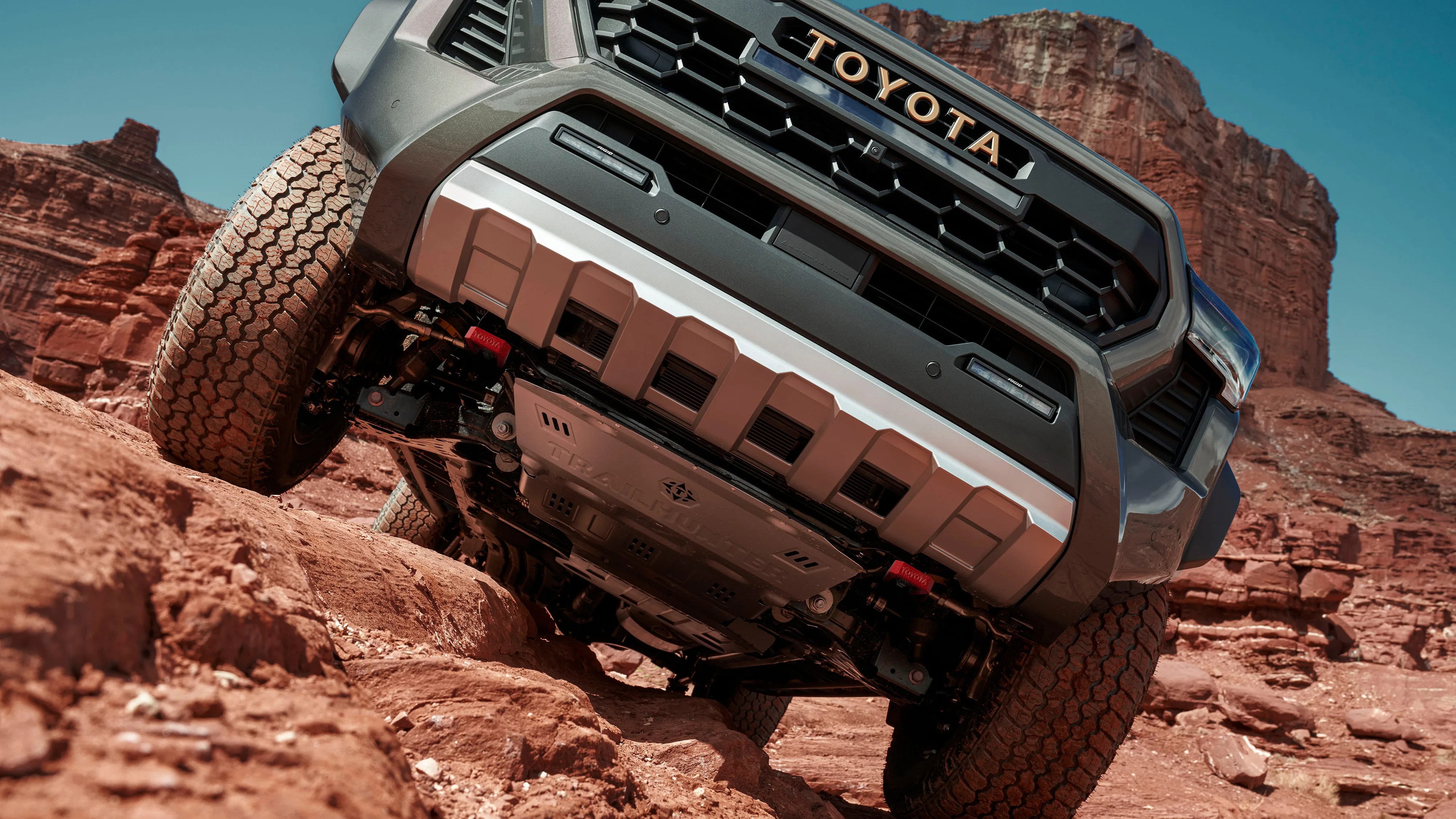 Front underside view of a gray Toyota truck navigating rocky red terrain with large tires and skid plates.
