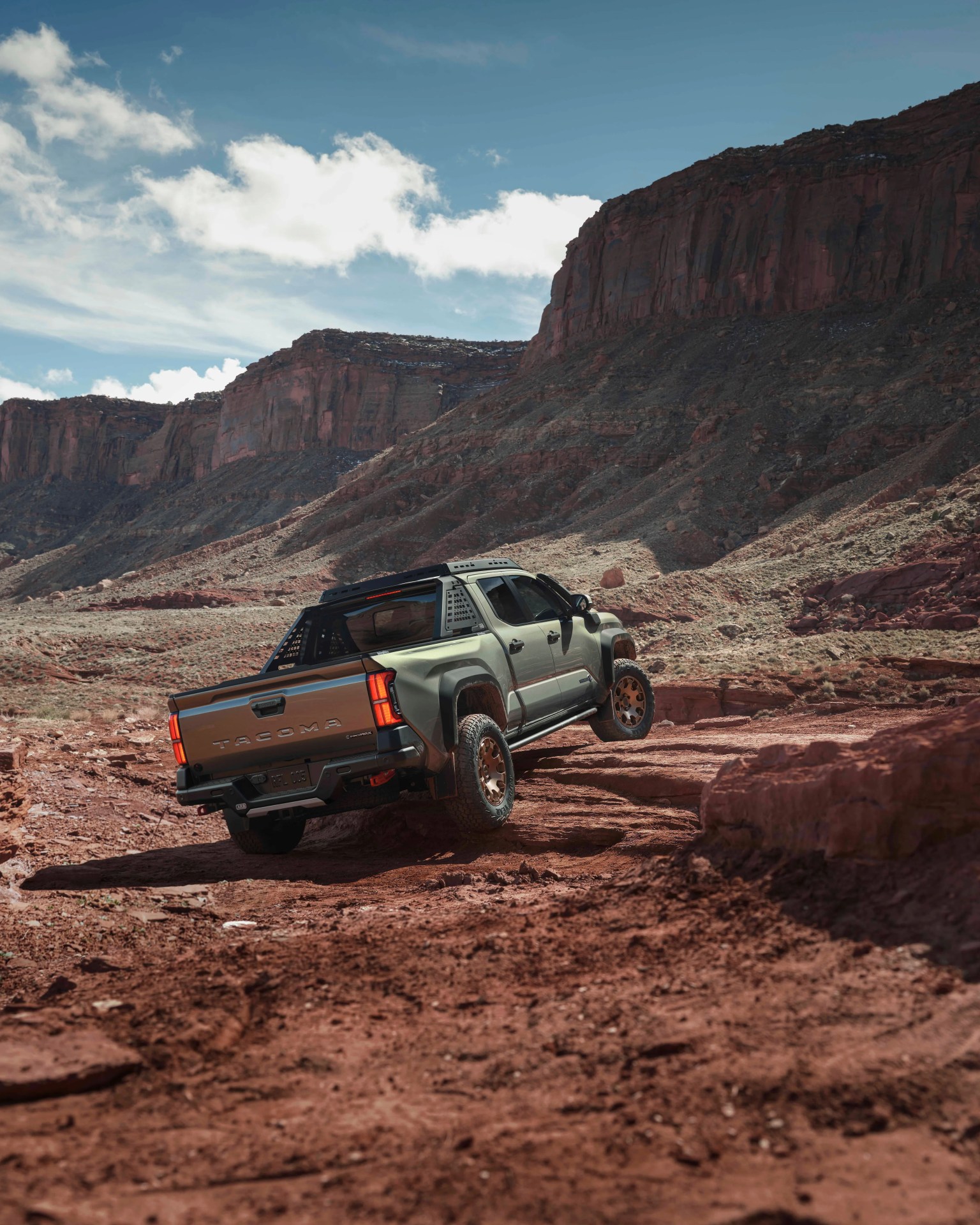 Green Toyota Tacoma pickup truck driving on rocky red dirt terrain with large cliffs in the background.