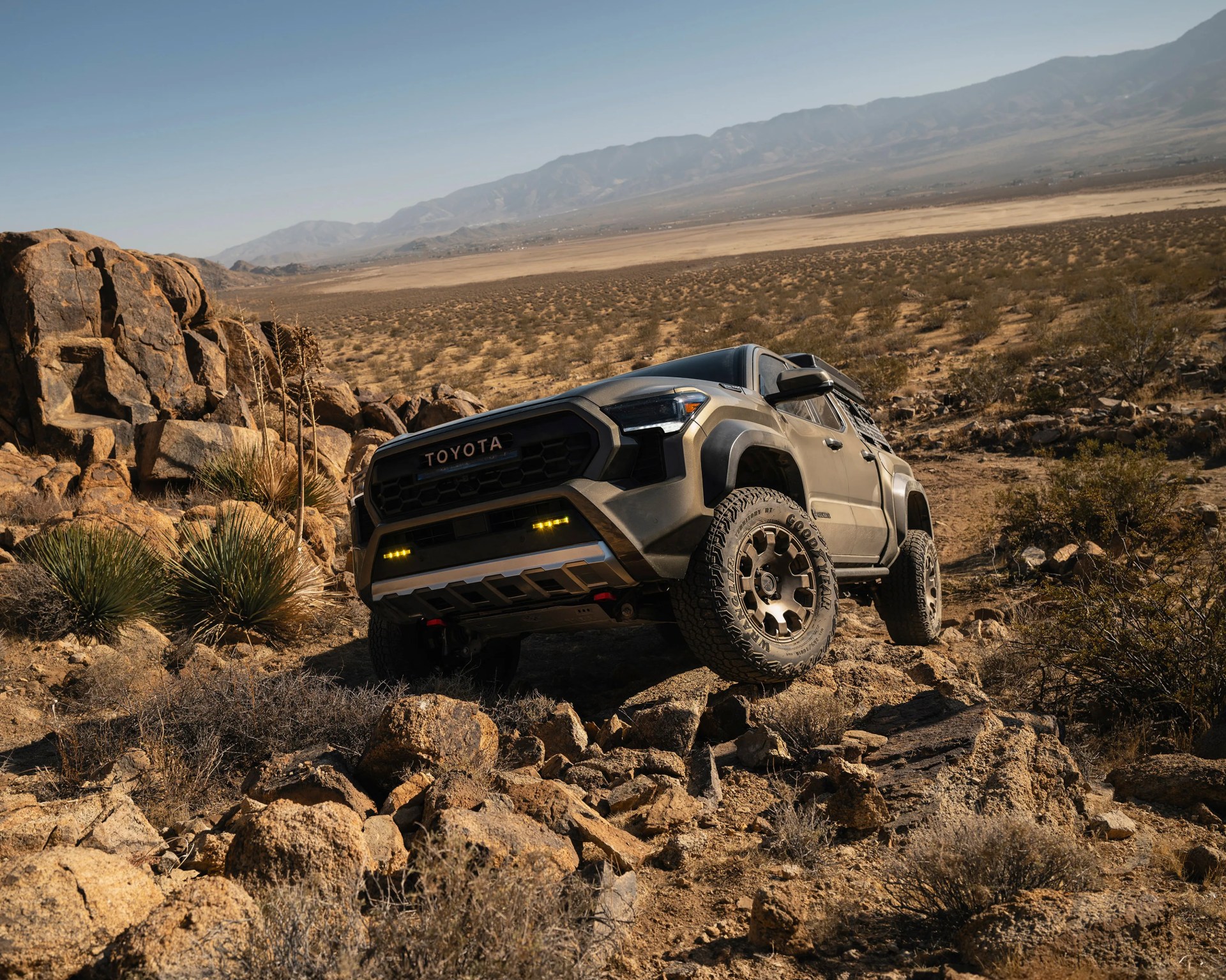 Matte gray Toyota off-road truck climbing rocky desert terrain with mountains in the background.