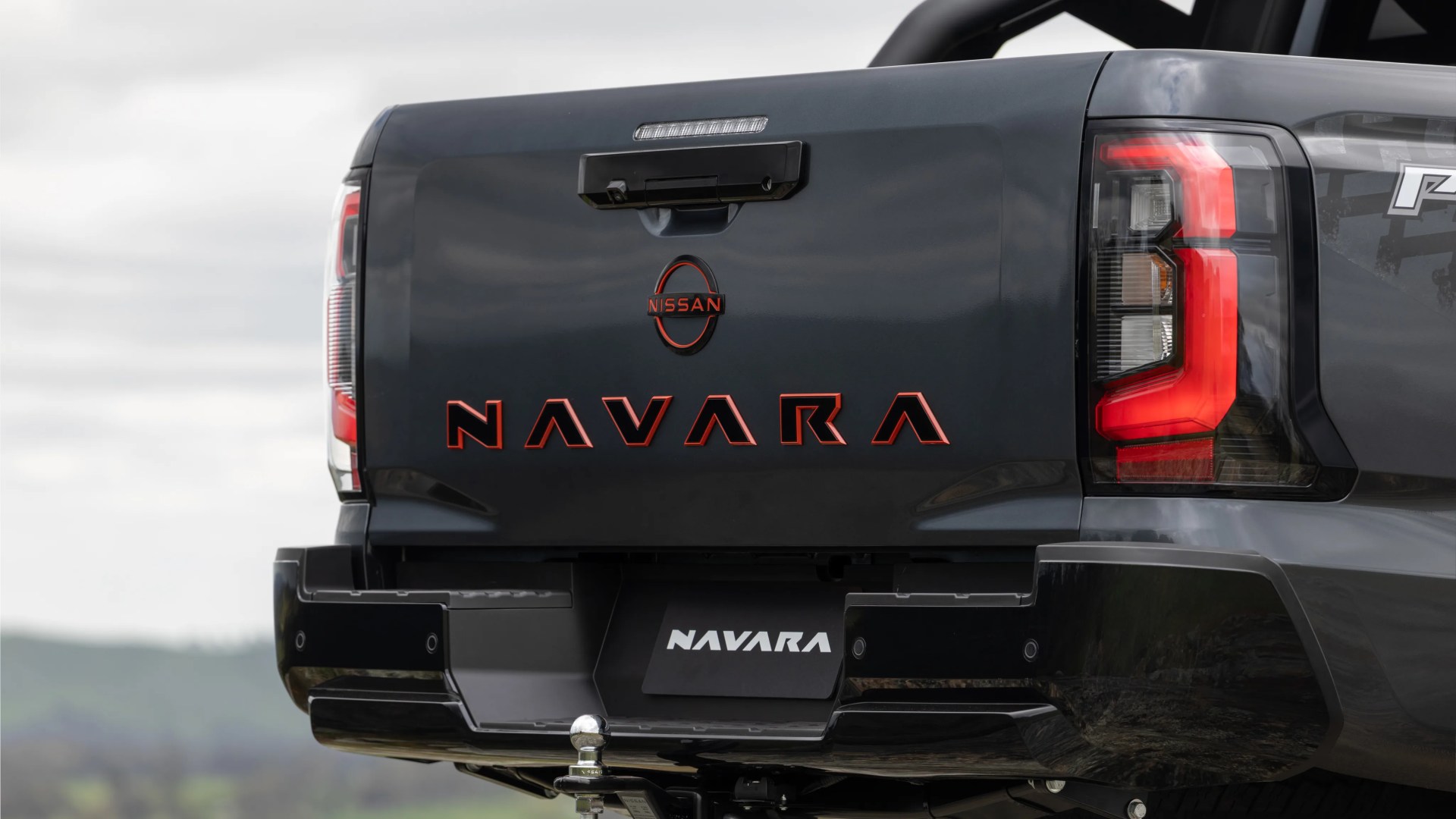 Rear view of a dark gray Nissan Navara pickup truck tailgate with red illuminated taillights and a tow hitch.