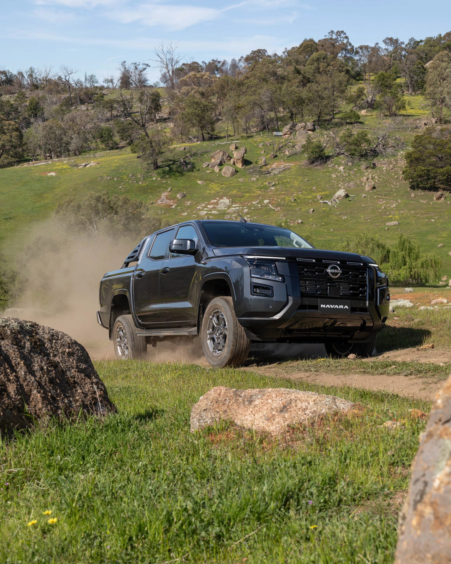 Black Nissan Navara pickup truck kicking up dust on a dirt trail in a green hilly landscape.