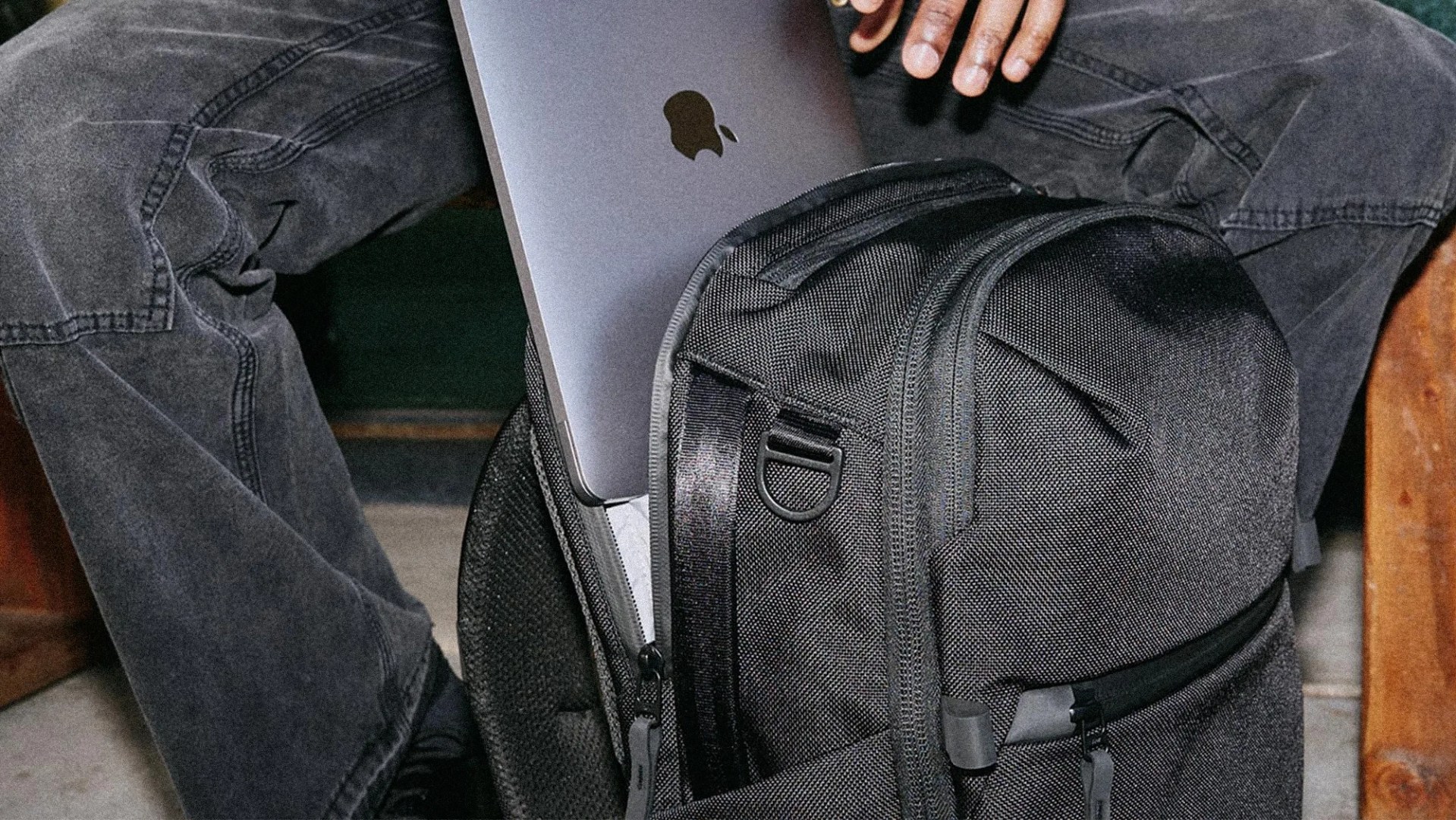 Black textured fabric backpack with a laptop partially inserted in the main compartment, worn by a person in dark jeans.